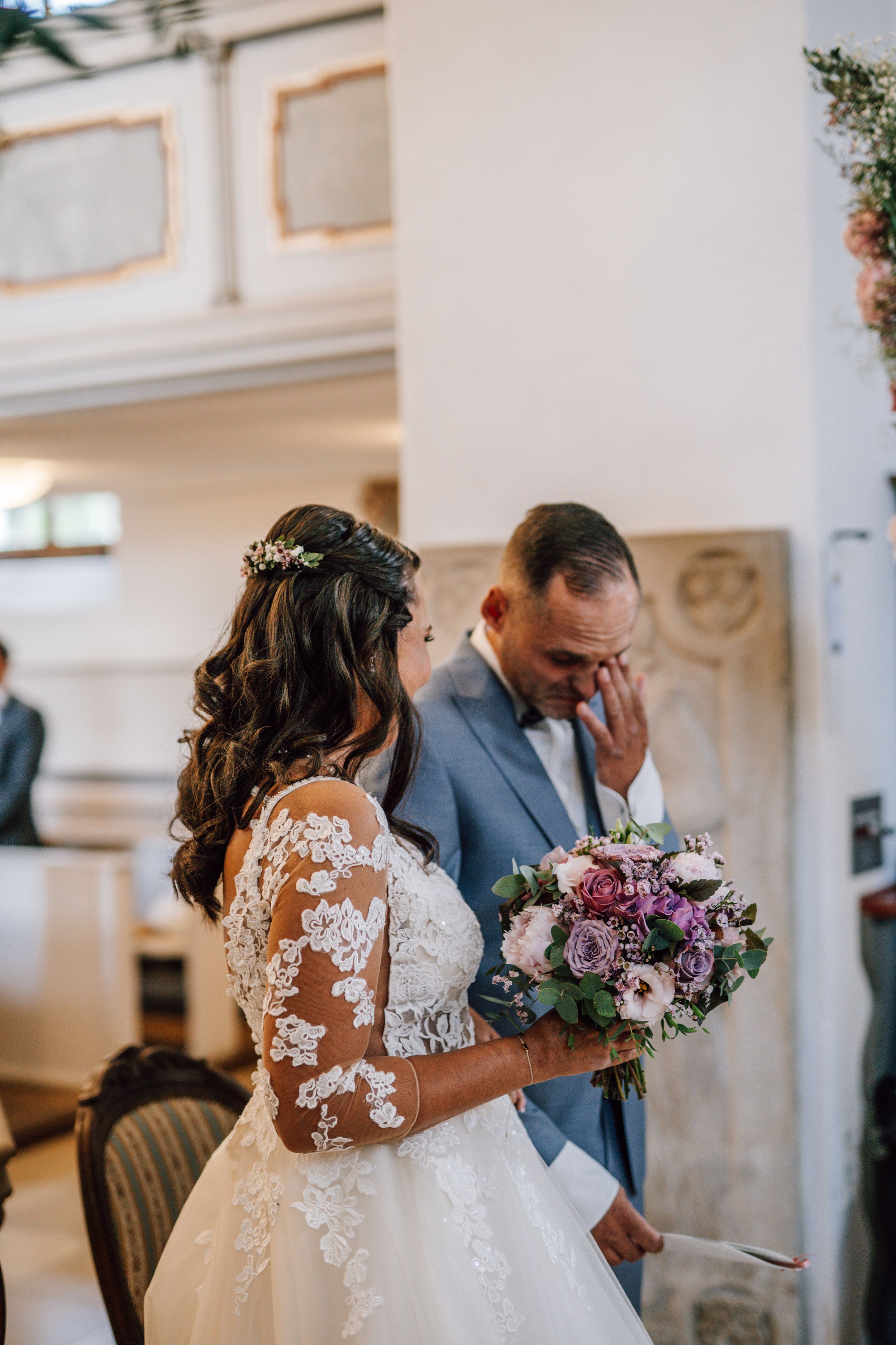 Wedding couple sharing an emotional moment in an elegant white room with purple floral bouquet.