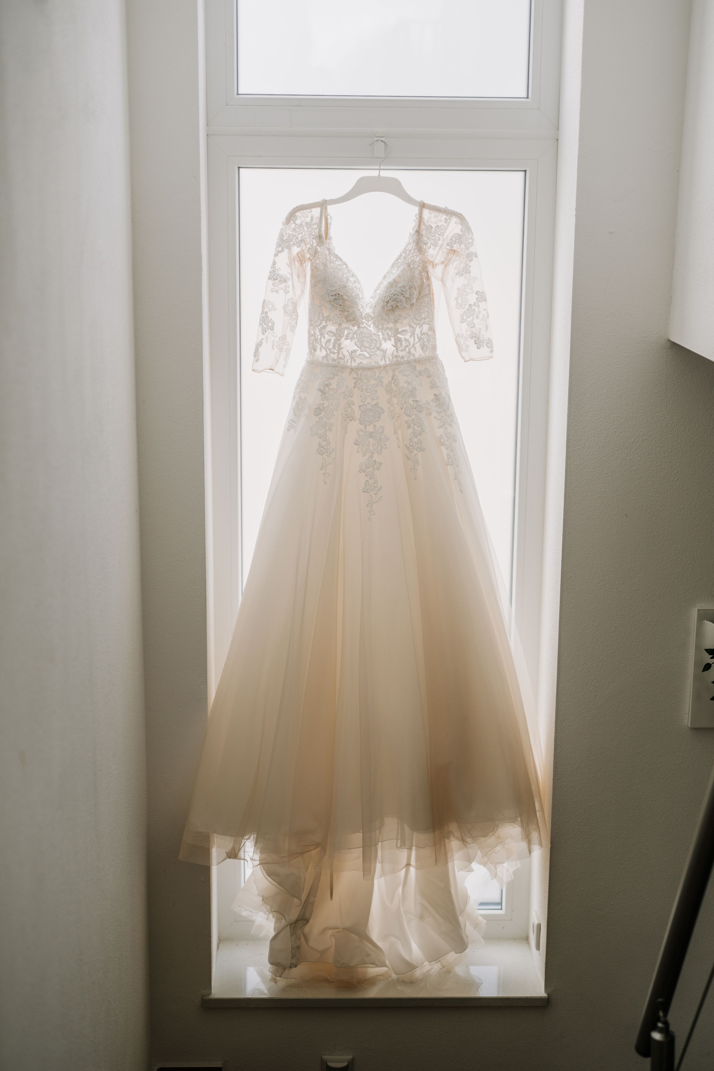 Ethereal backlit wedding dress hanging in window with soft natural light.