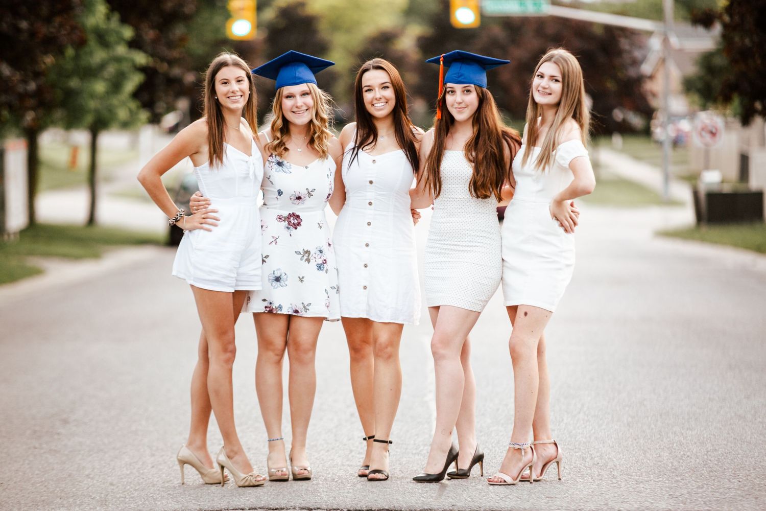 Five female graduates in white dresses and blue caps pose together on a street for a graduation photo.