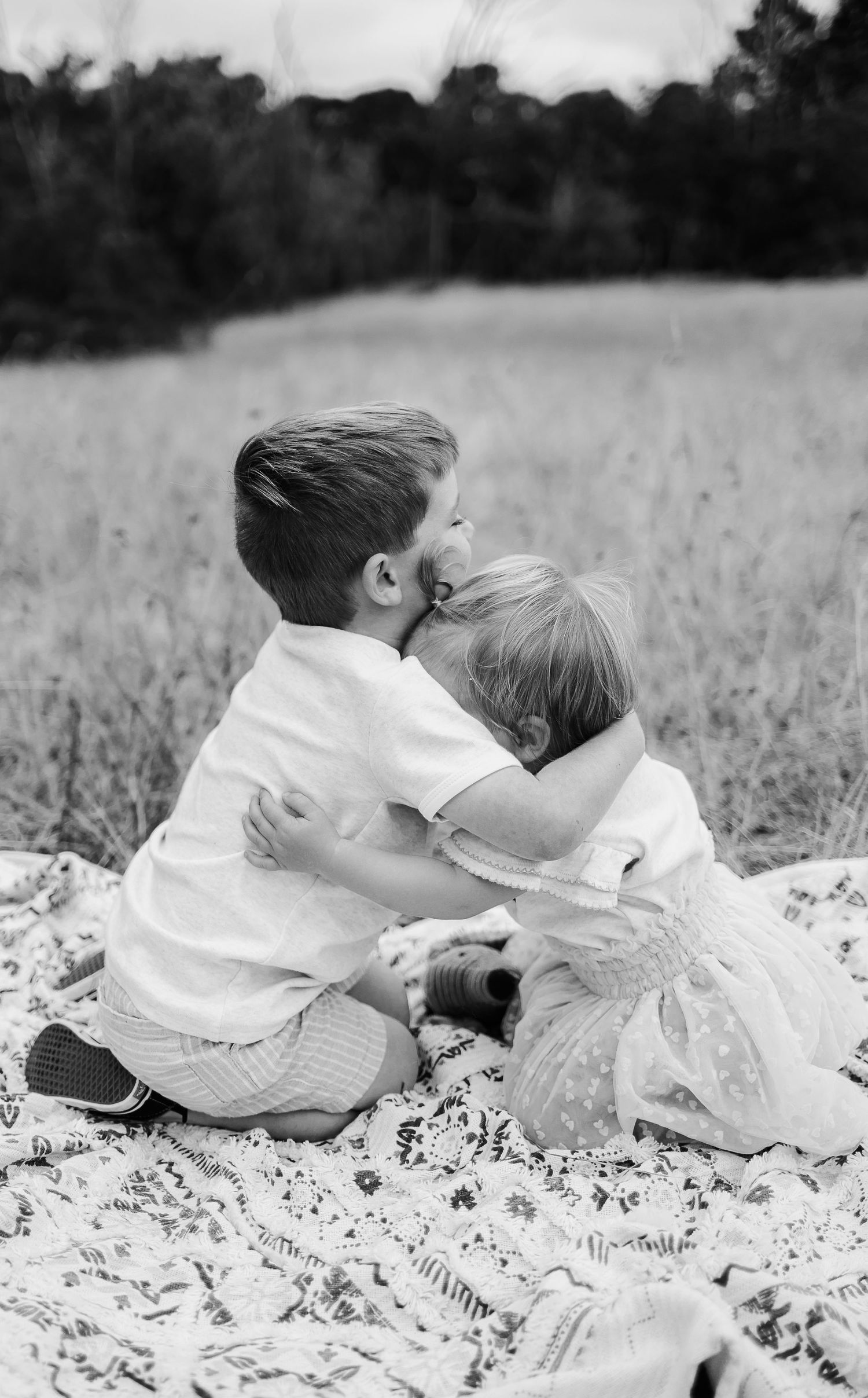 Two children share a tender embrace while sitting on a blanket in a grassy field.