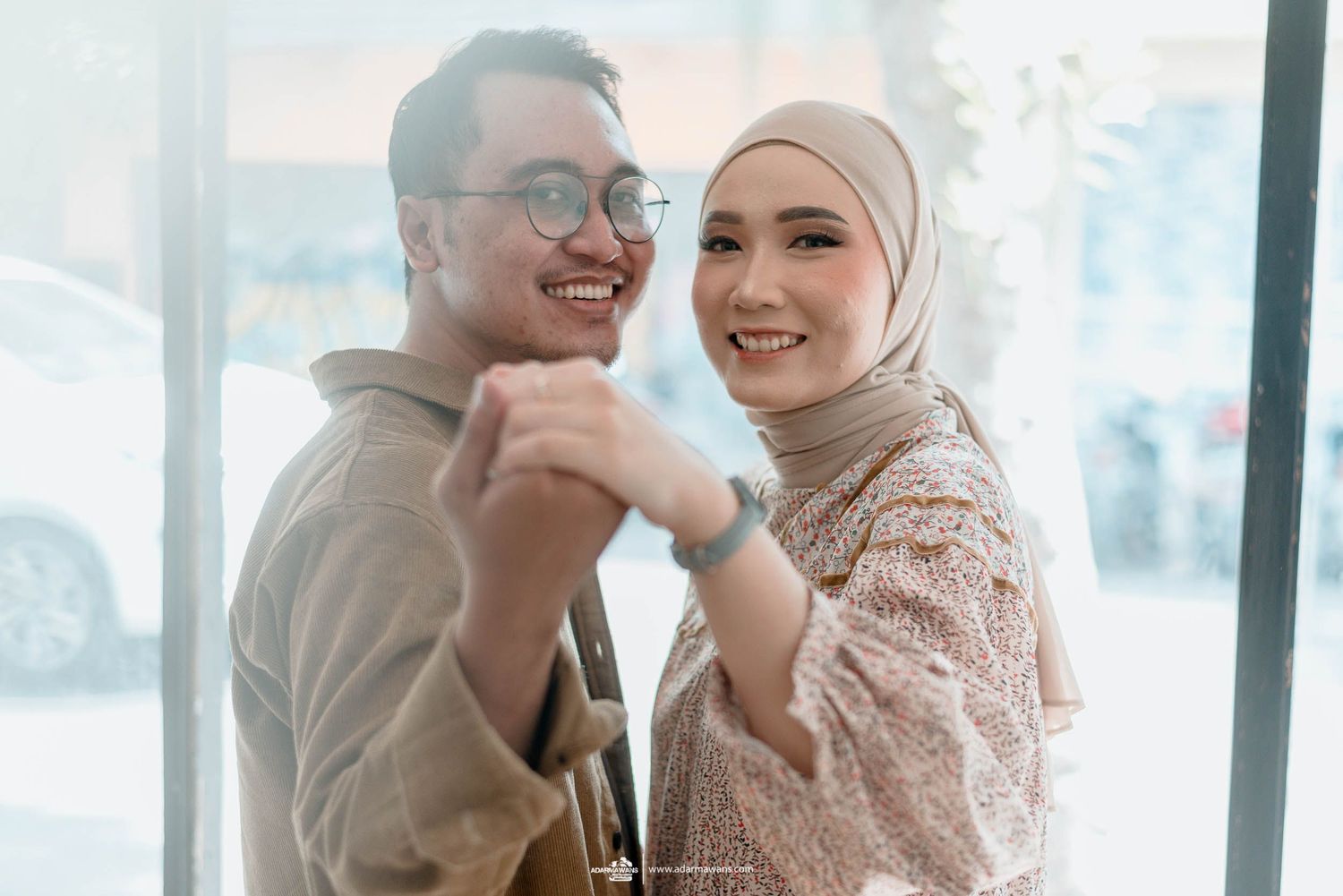 A joyful couple smiles while holding hands in front of a bright window.
