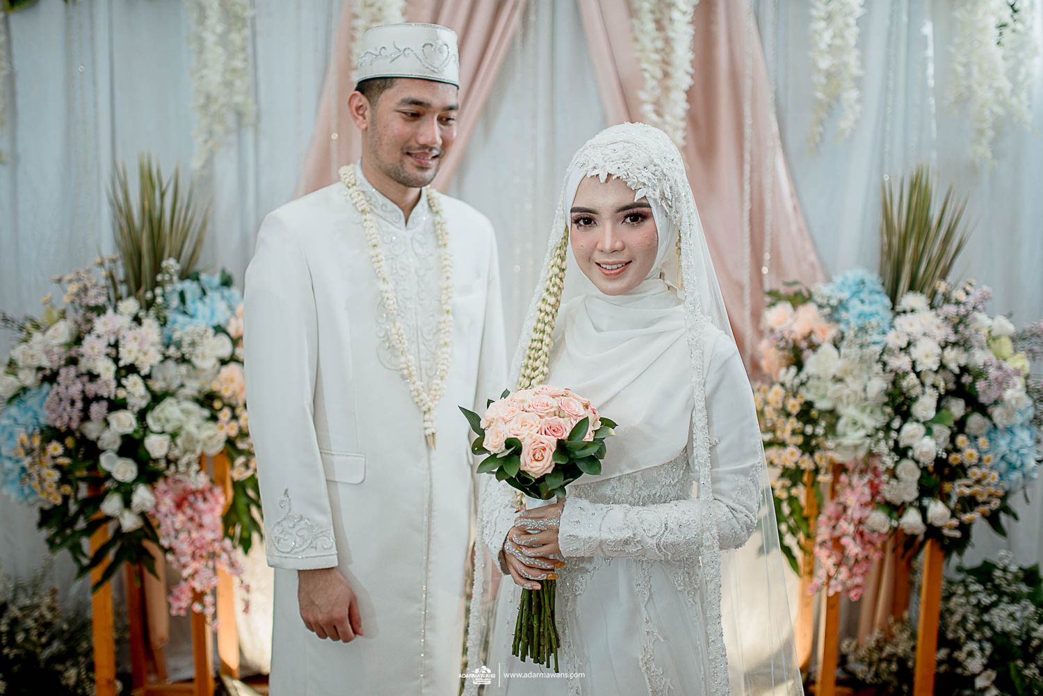 Muslim couple in traditional white wedding attire standing against floral backdrop.