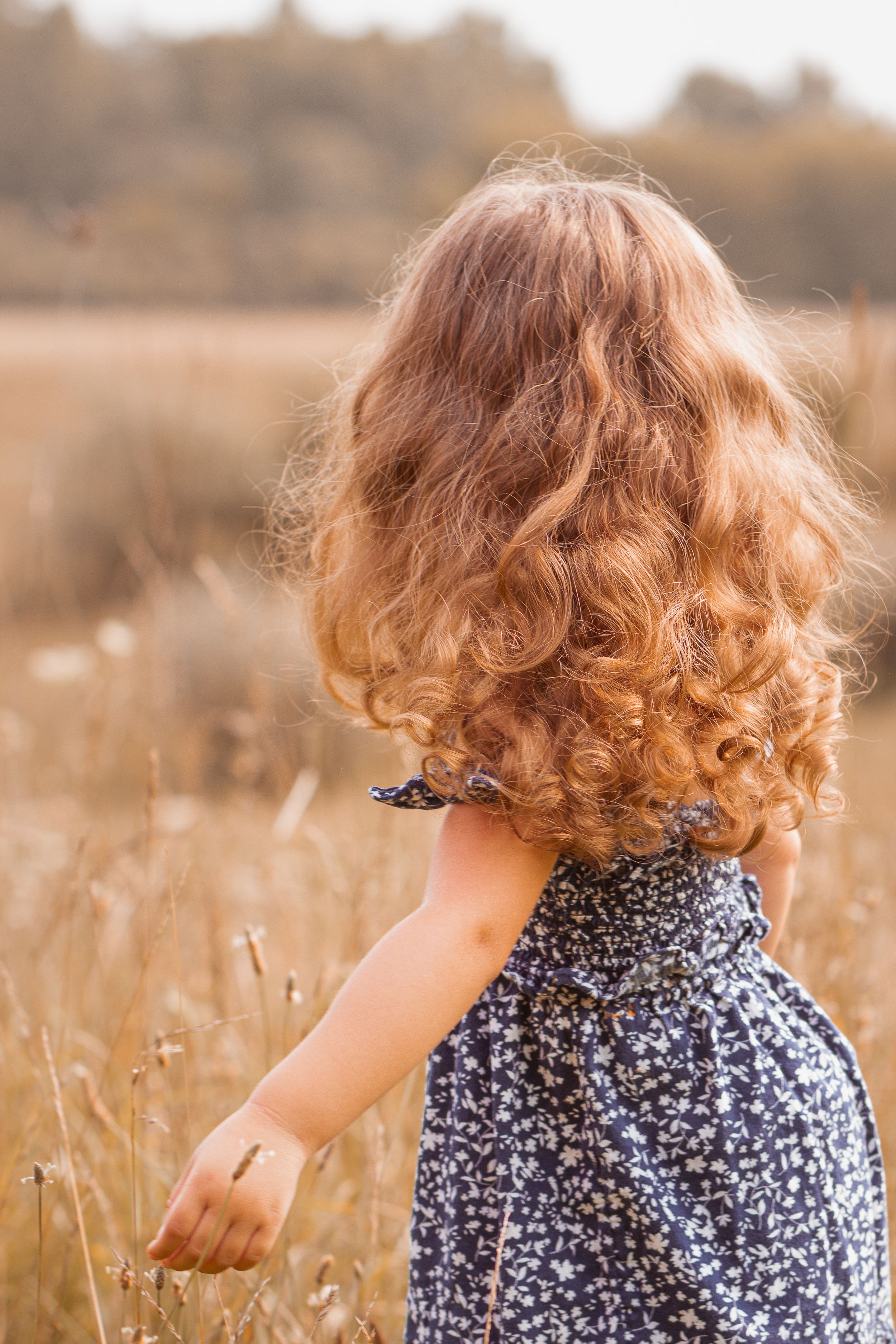 Child with curly ginger hair twirls in floral dress among tall grass.