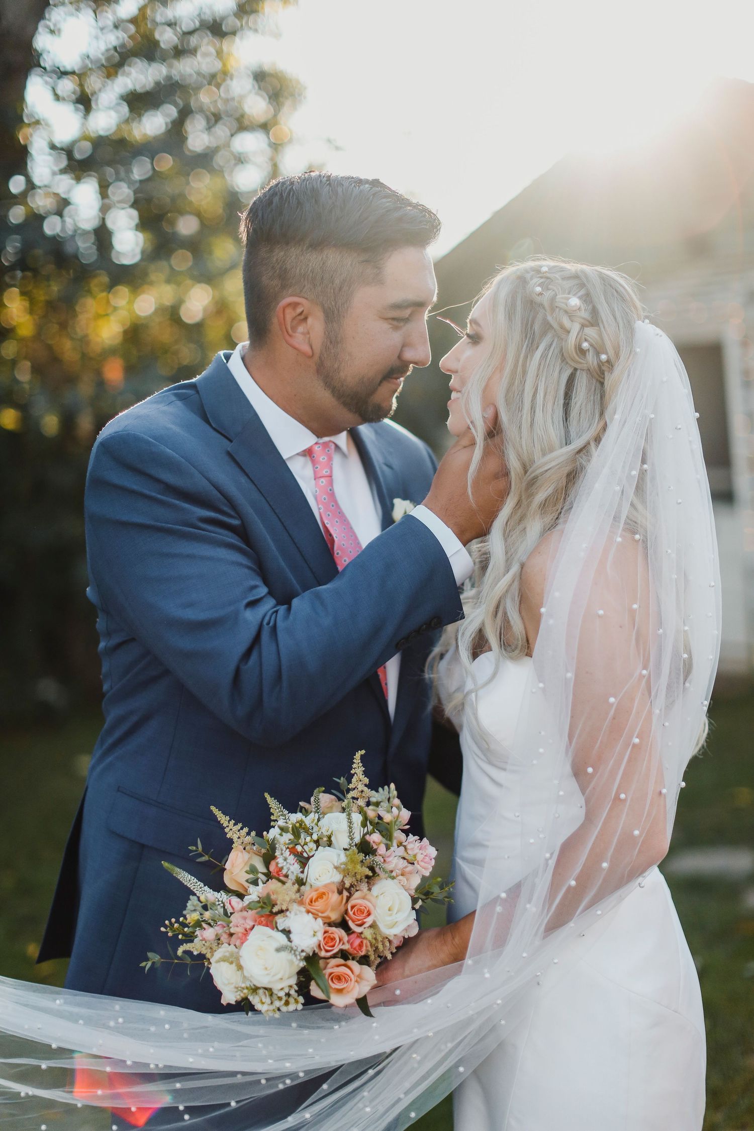 Romantic wedding couple shares intimate moment in sunlit garden with bride holding peach and white floral bouquet.