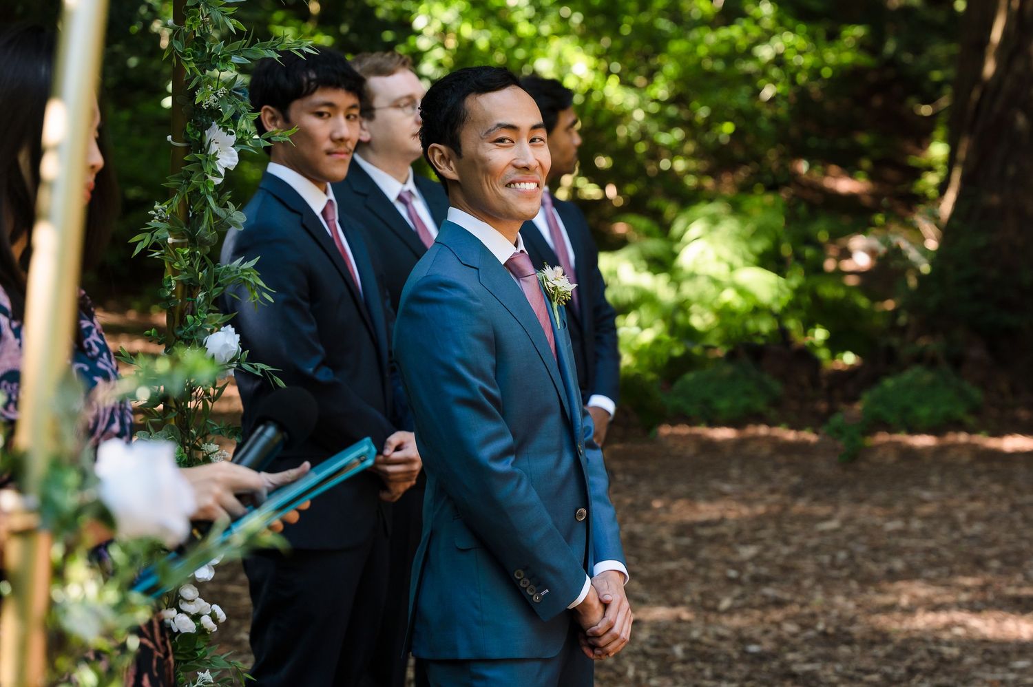 Groomsmen in navy blue suits stand during an outdoor forest wedding ceremony.