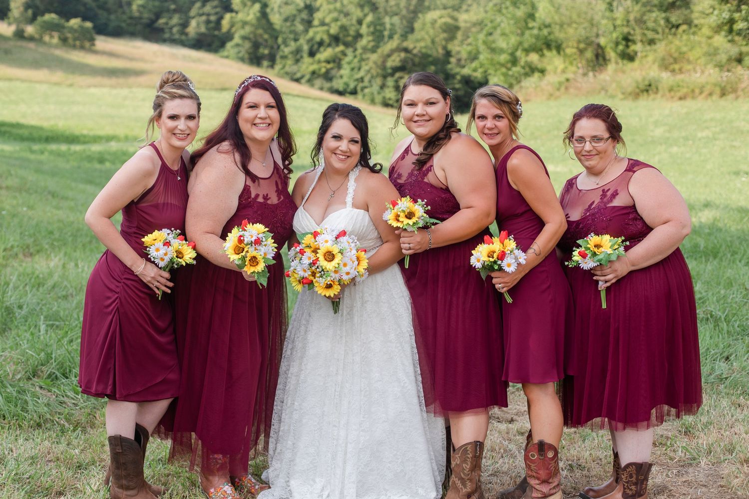 Wedding party in burgundy dresses holding vibrant sunflower bouquets pose together in a grassy field.