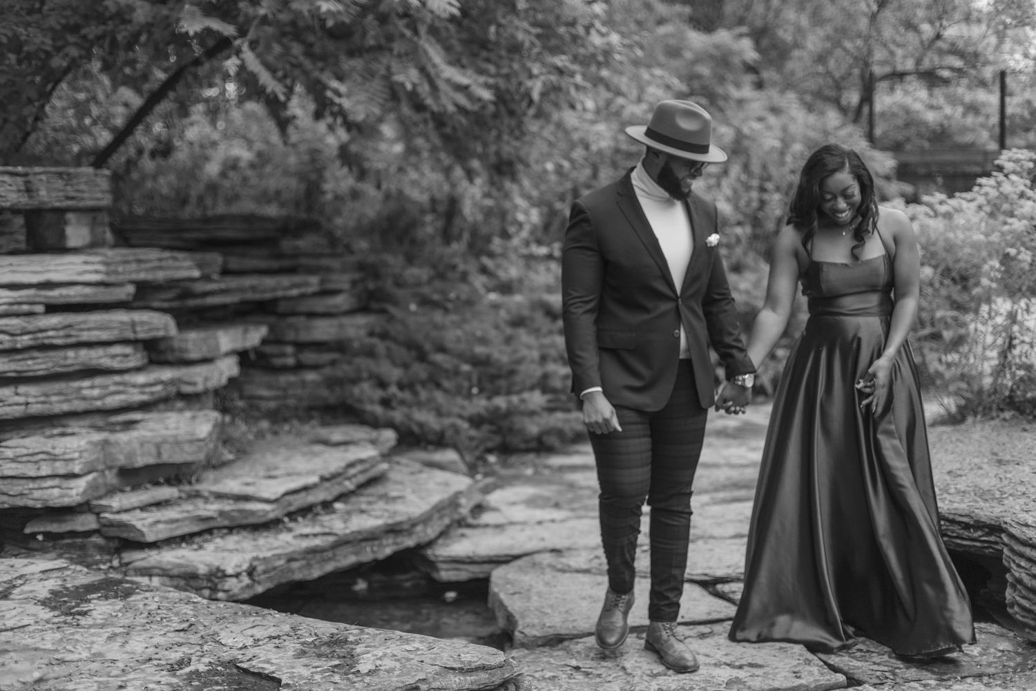 A couple in formal attire poses together on stone steps near a waterfall in black and white.