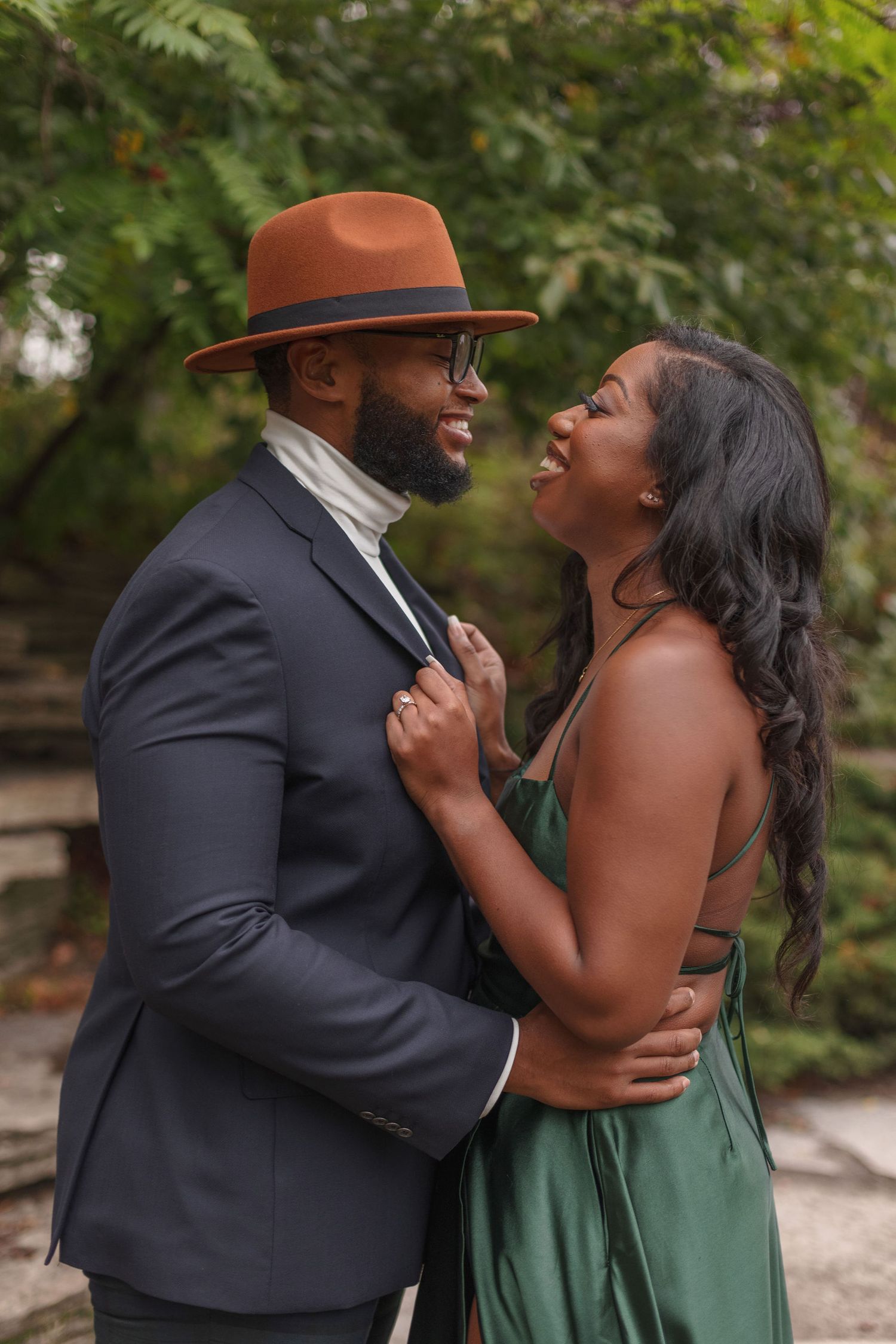 A romantic couple embraces outdoors, with one wearing a brown fedora and navy suit.
