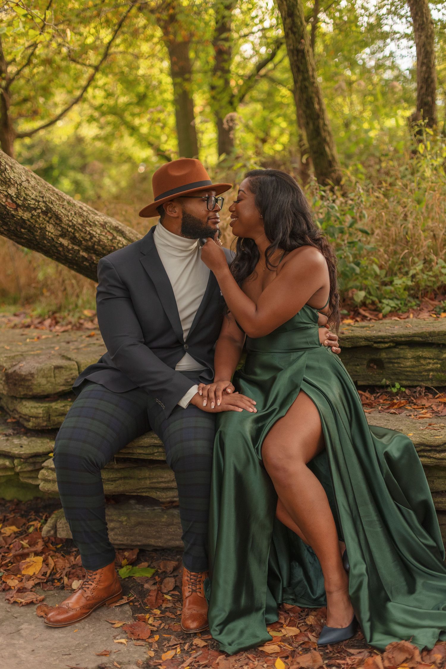 Romantic couple in elegant attire sitting on stone wall in autumn park setting.