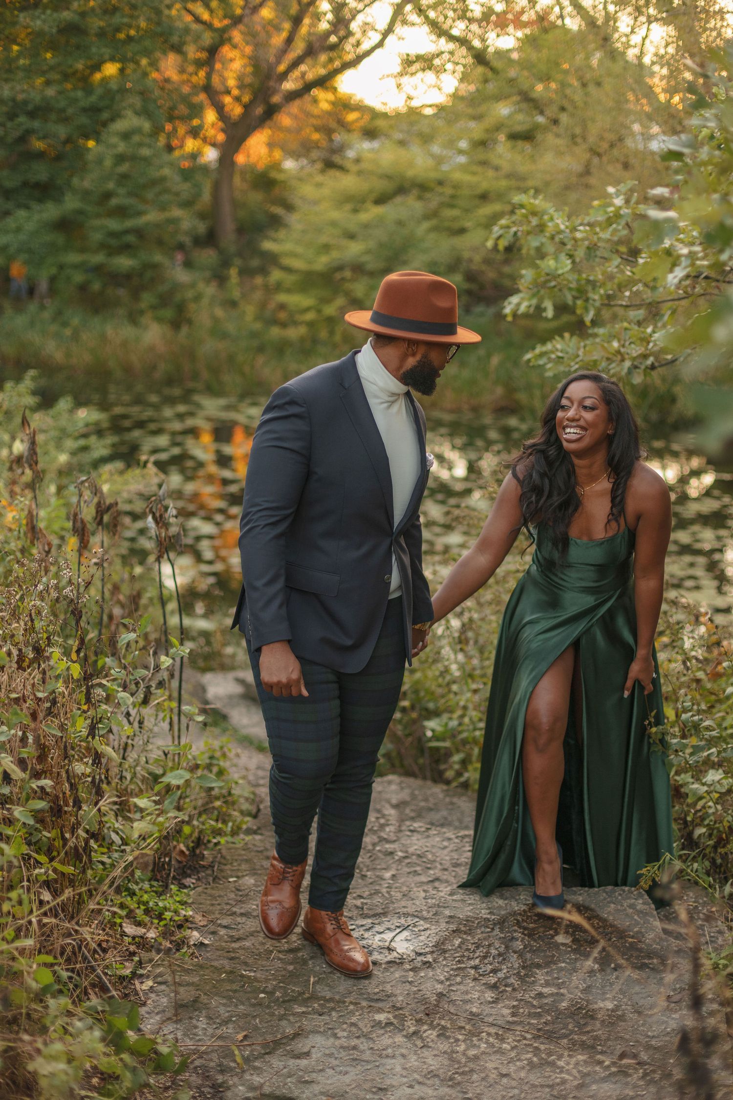 A couple walks along a nature trail during golden hour, one wearing an elegant green dress.