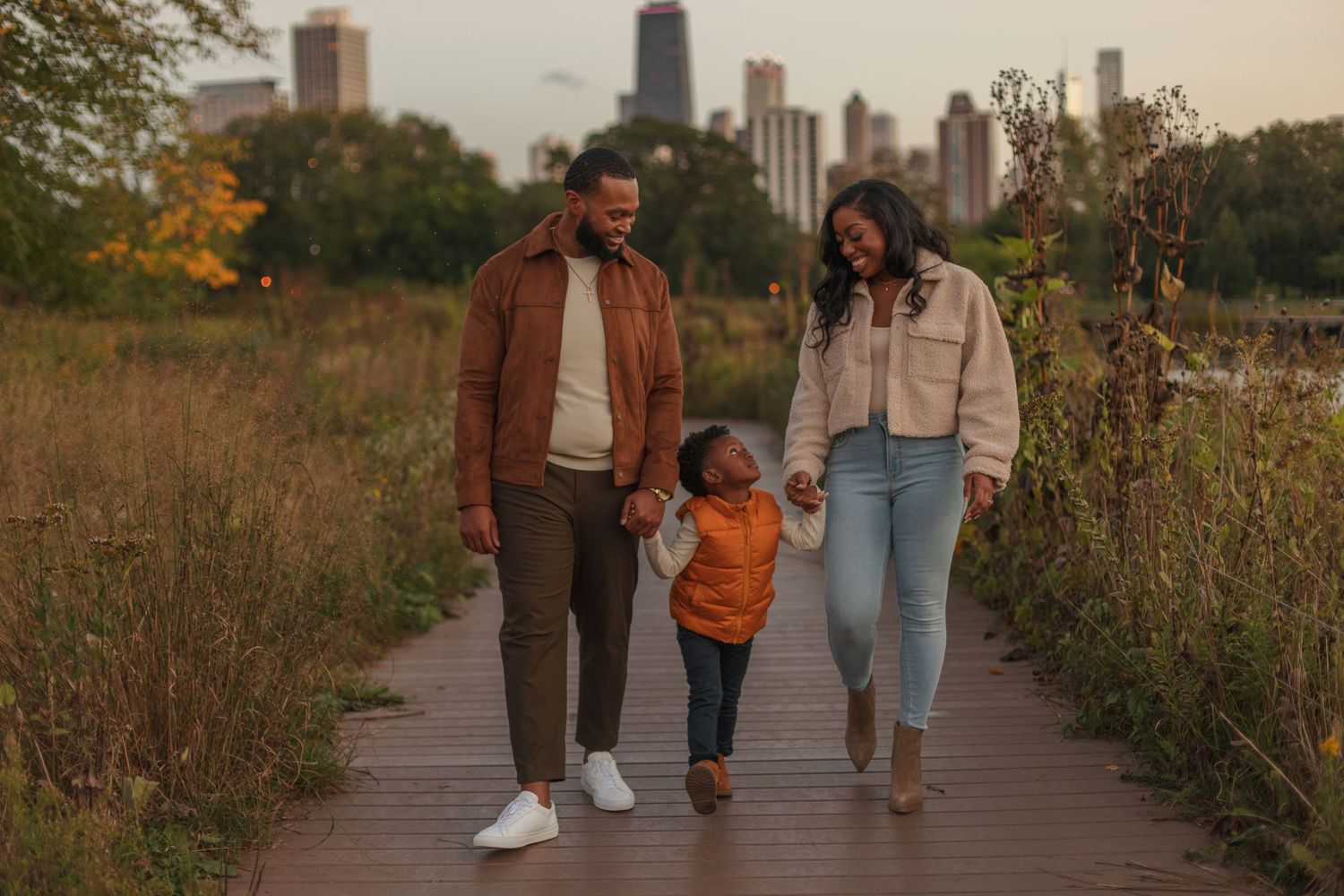 Family walking together on a wooden path through tall grass with city skyline in background at sunset.