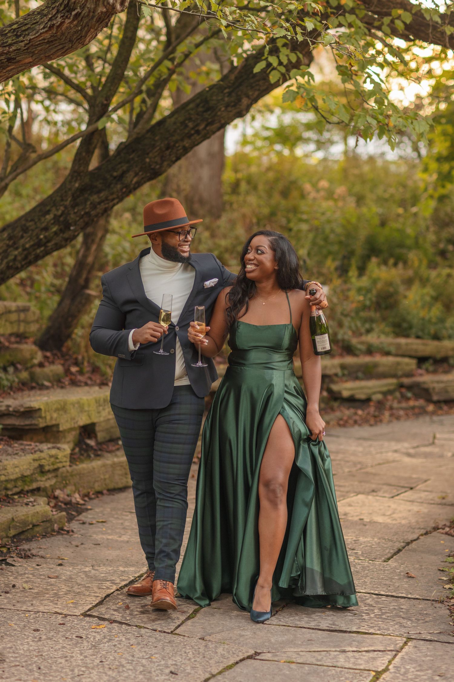 Stylish couple in formal attire sharing drinks while walking on stone path in garden at sunset.
