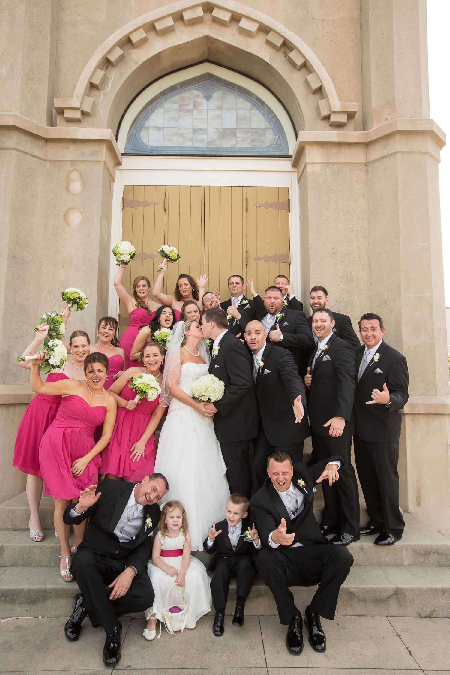 Wedding party in pink dresses and black suits posing together outside church.