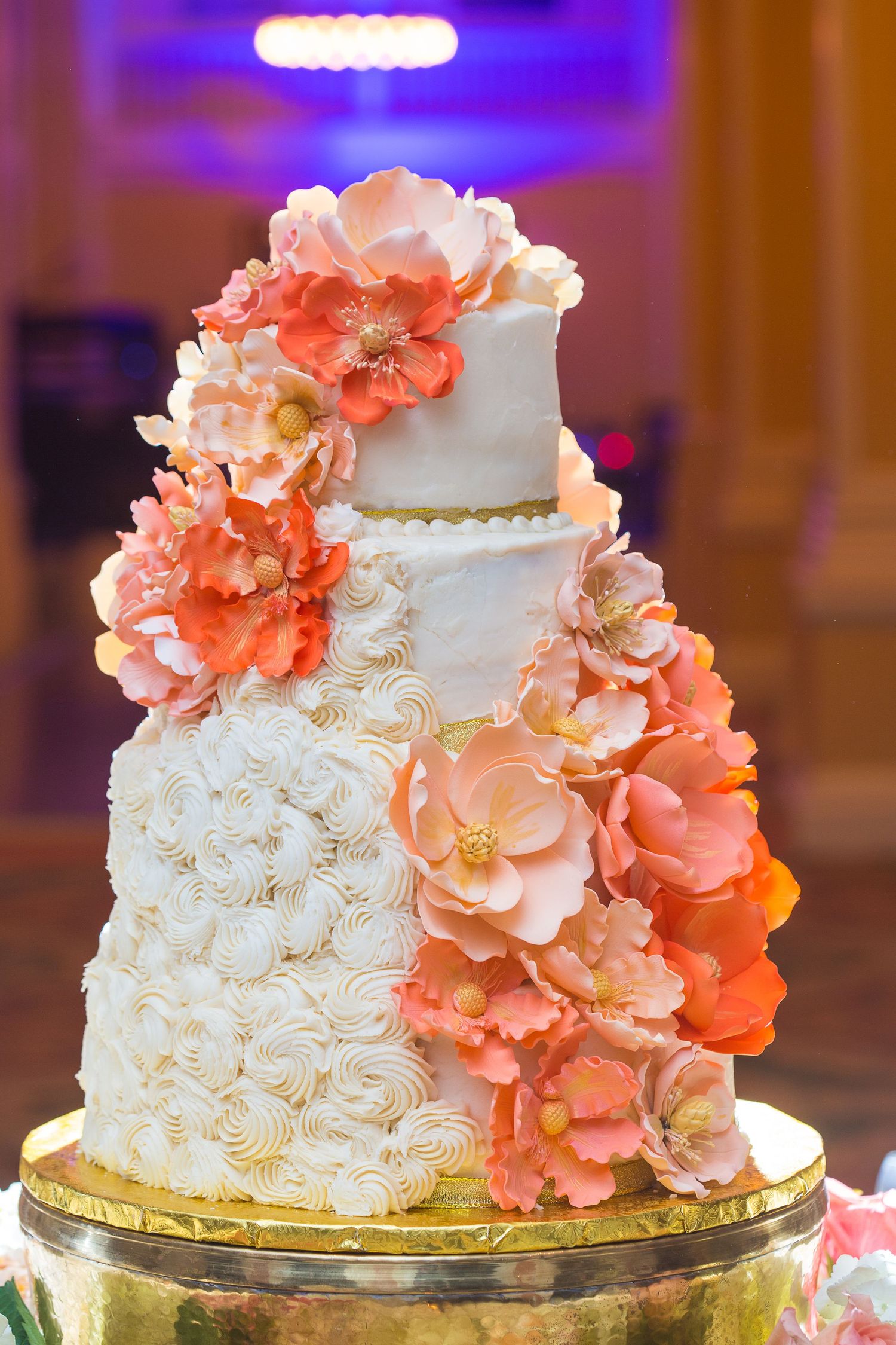 Elegant white wedding cake decorated with coral-colored sugar flowers and gold accents on a metallic stand.