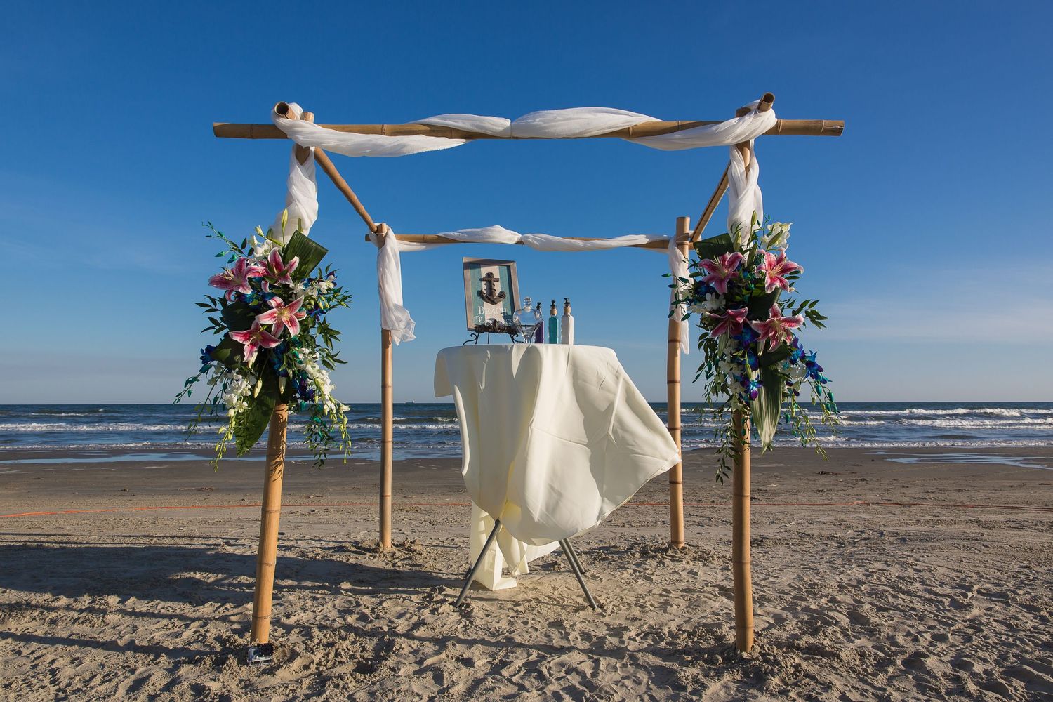 Wooden arch decorated with white fabric and flowers on a sandy beach.