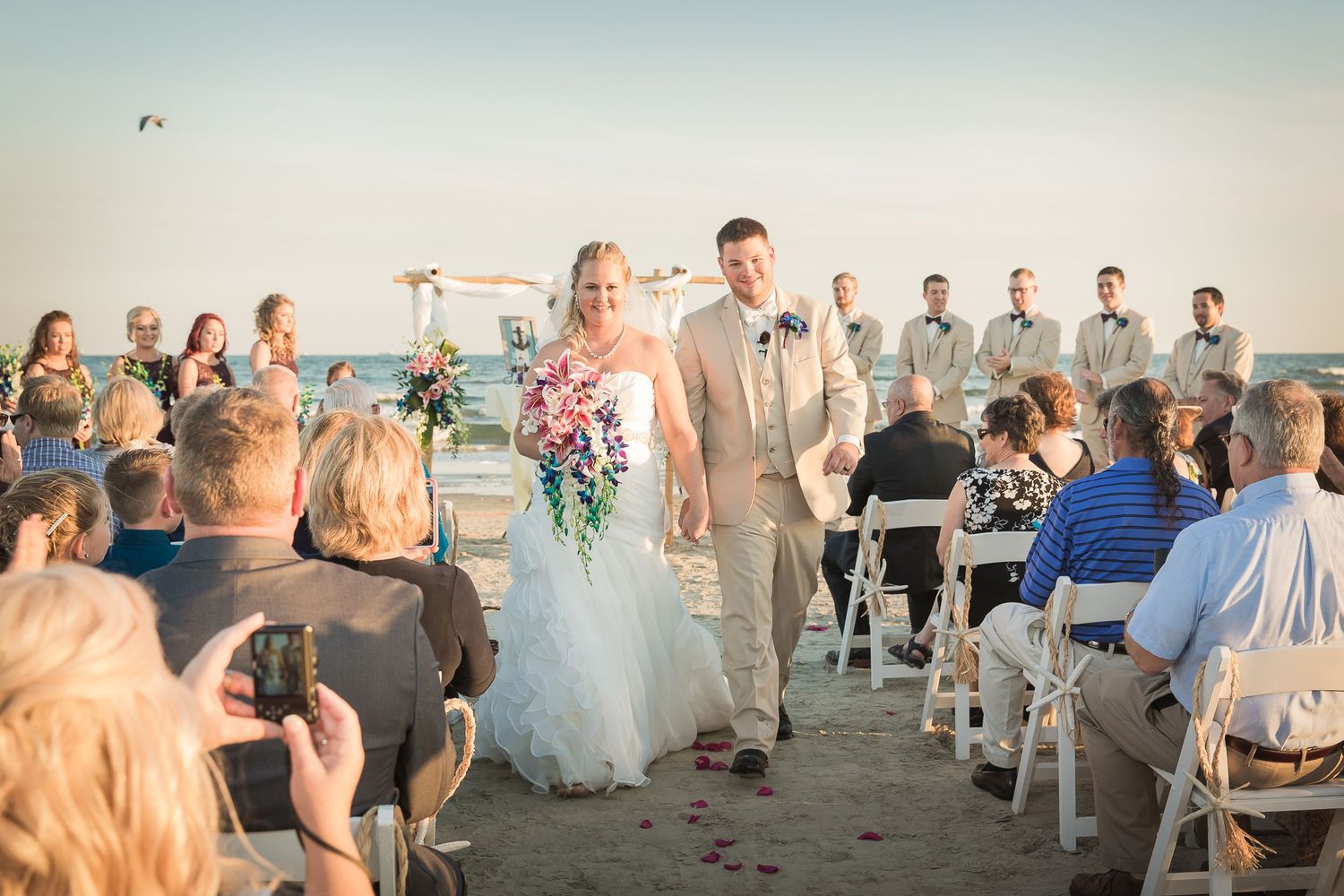 Beach wedding ceremony setup with white chairs and floral decorations at sunset.