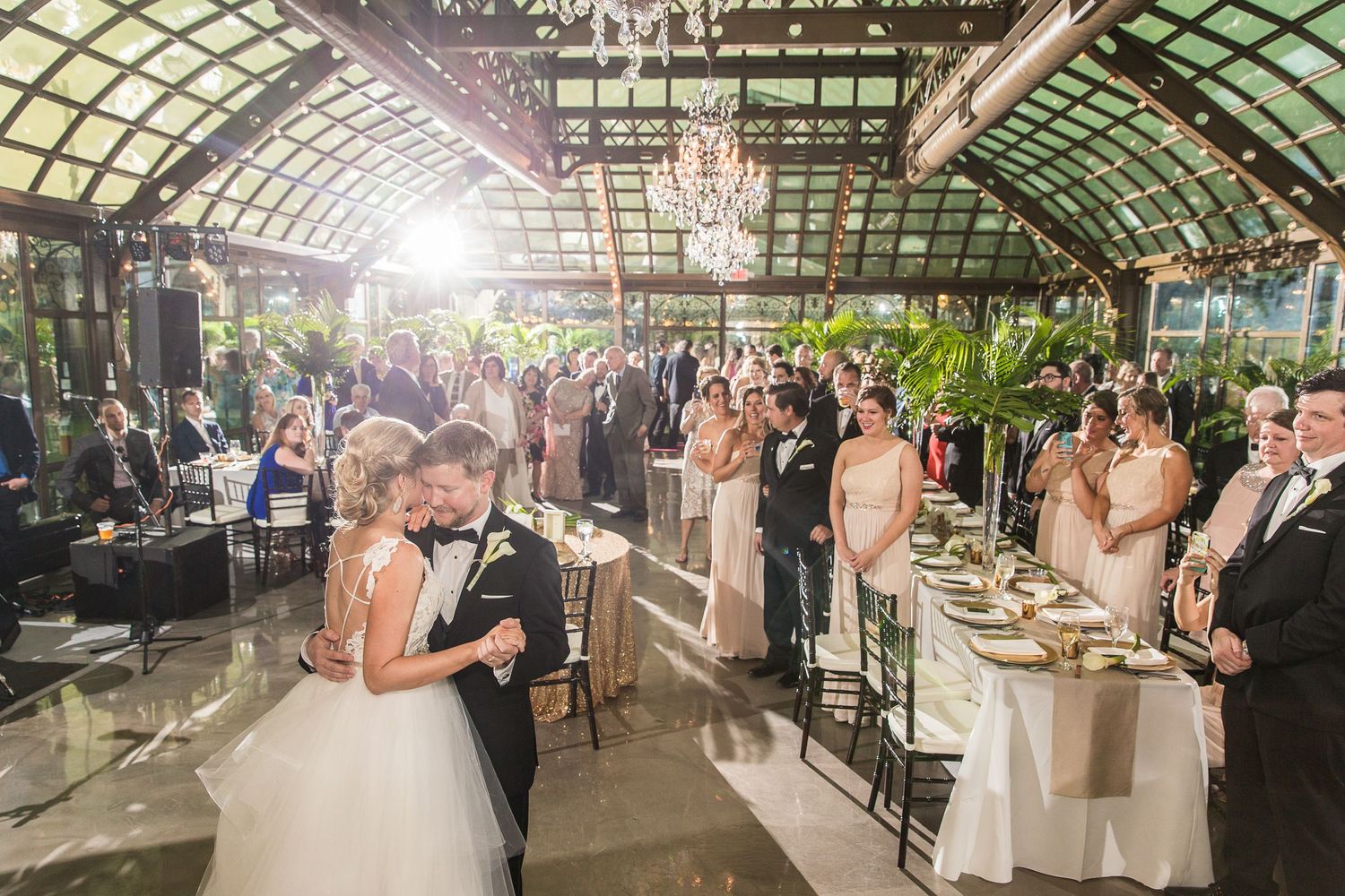 Wedding reception celebration in a greenhouse-style venue with chandeliers and natural lighting.