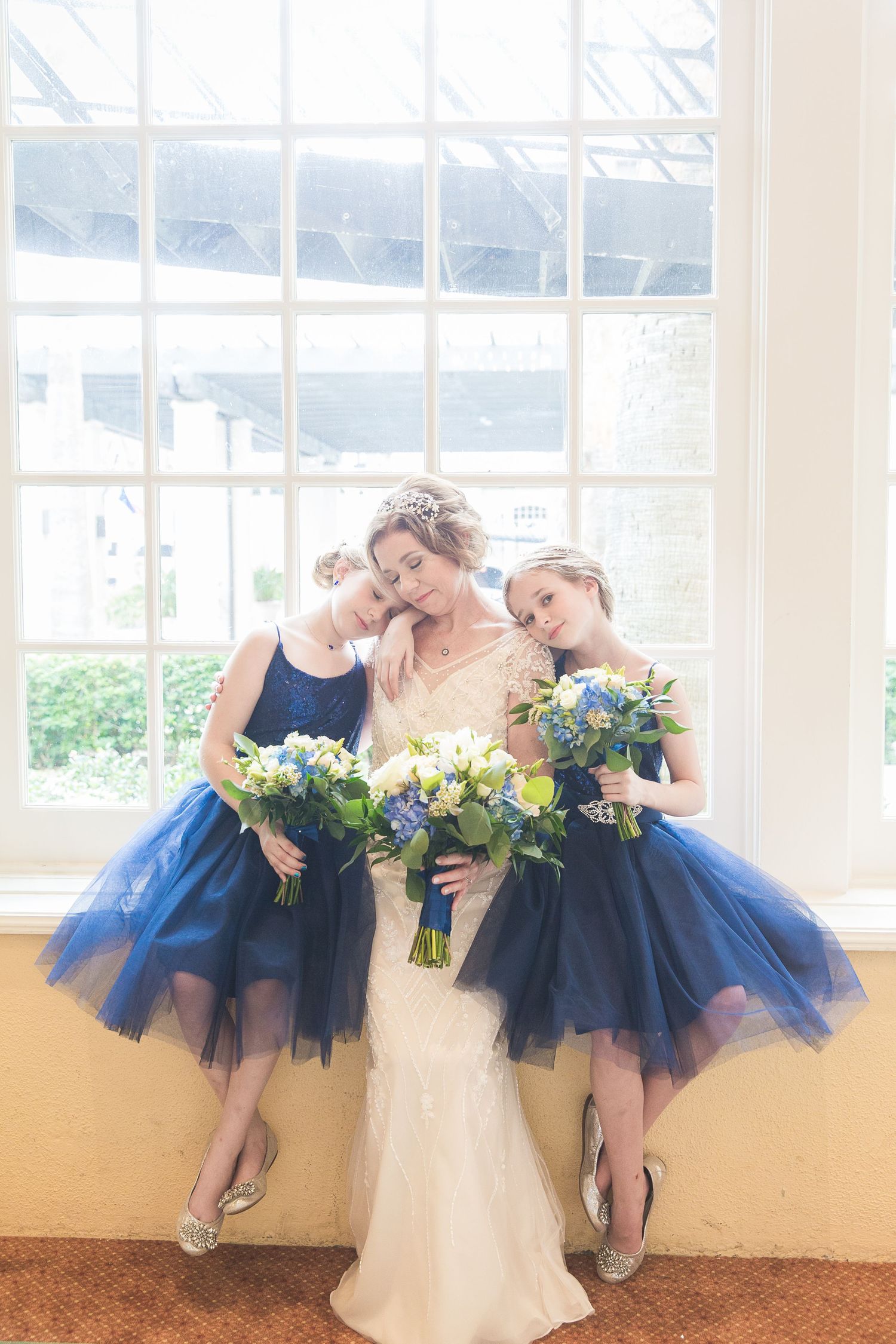 Bridal party in navy blue tulle dresses posing playfully by sunny windows.