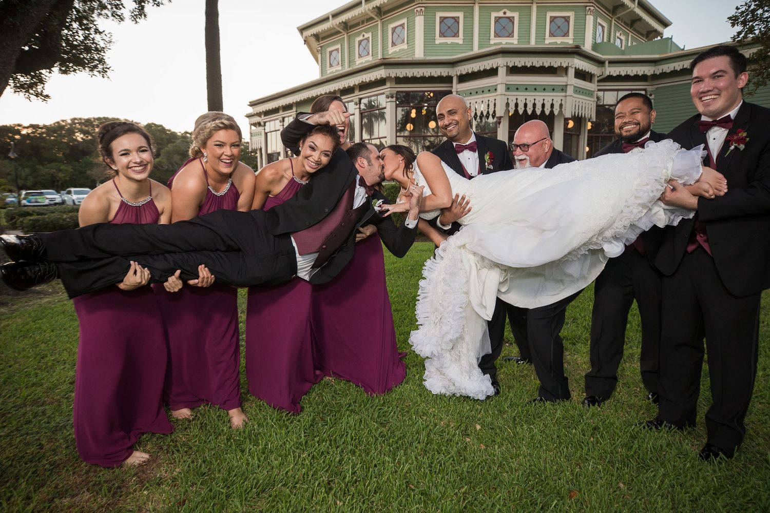 Wedding party posing playfully on grass with bride's veil flowing in the wind.