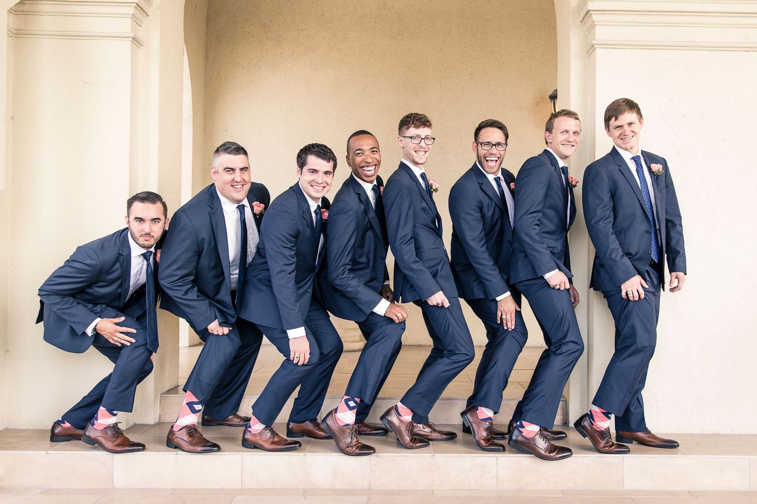 Groomsmen sitting in a row showing off matching brown shoes against a light colored wall.