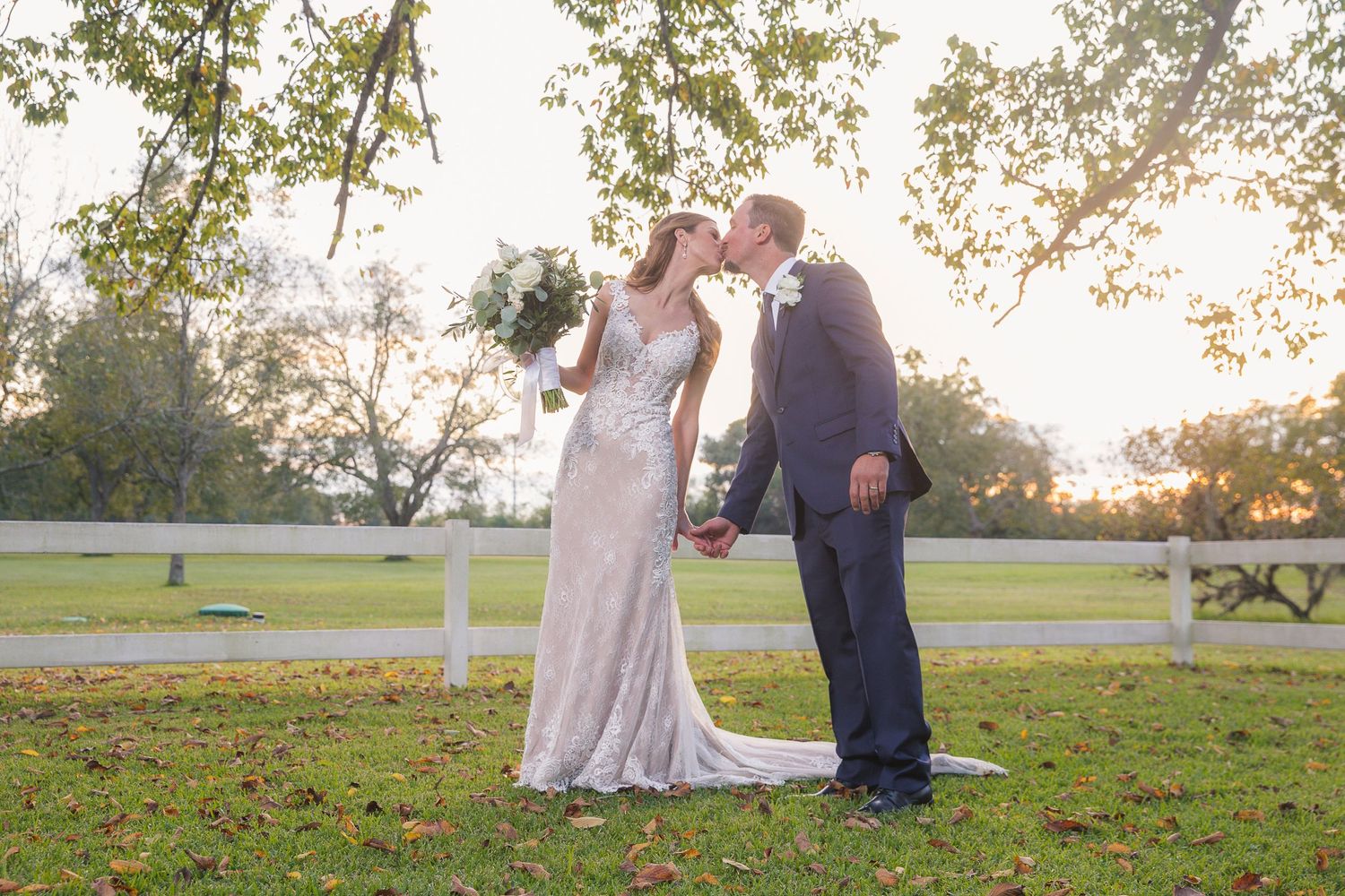 Couple embraces on a grassy hill during sunset with white fence in background.