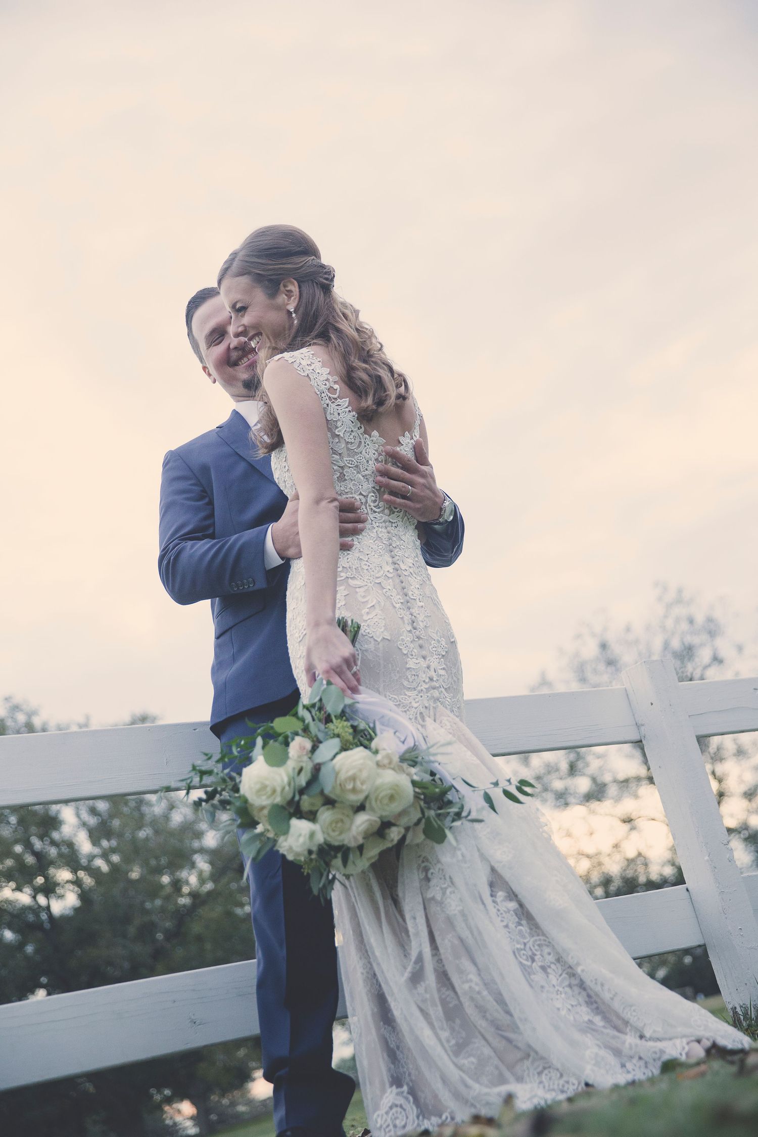 Romantic moment captured against white fence with bride holding elegant white bouquet.