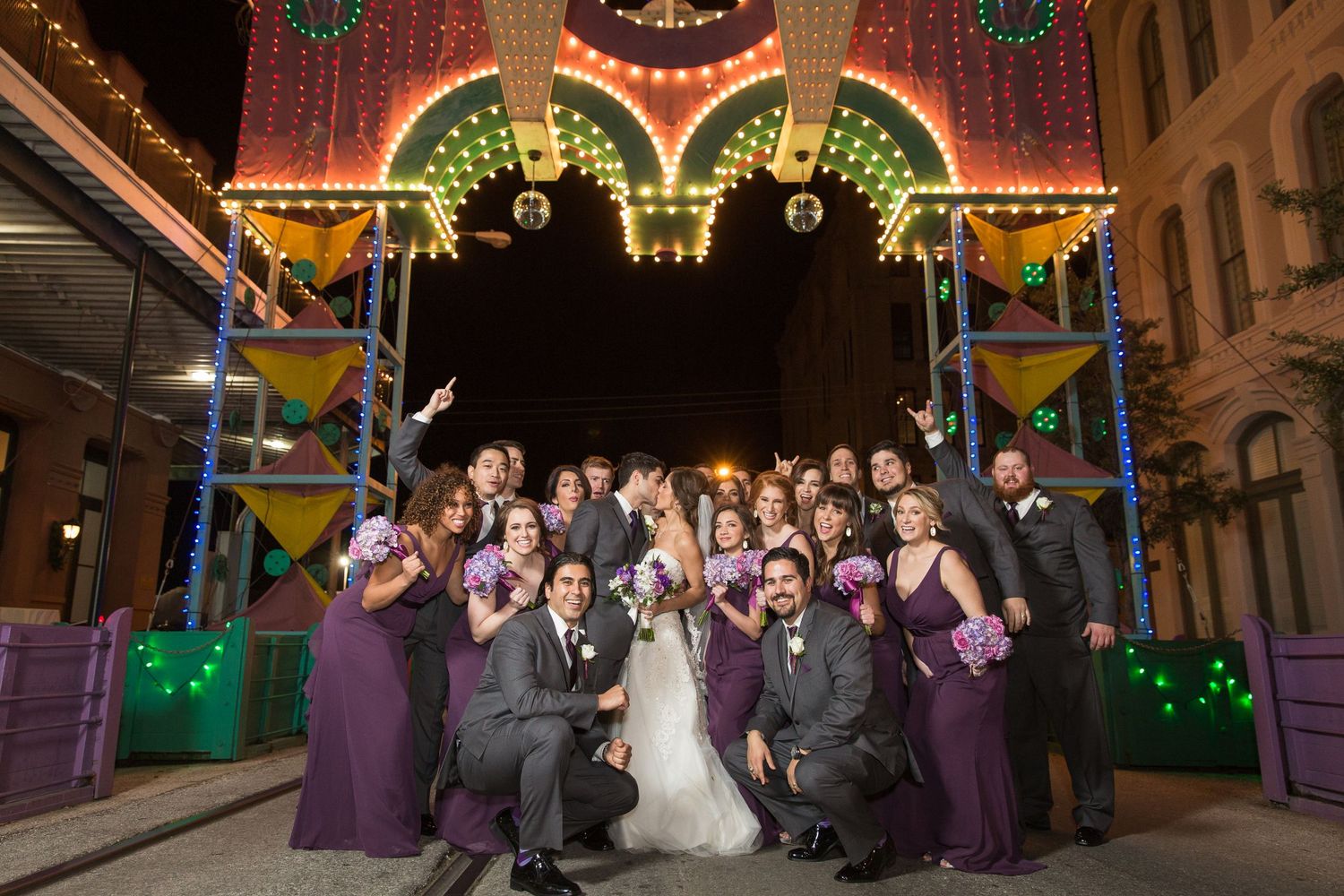Wedding party posing at night under festive street decorations with colorful lights.