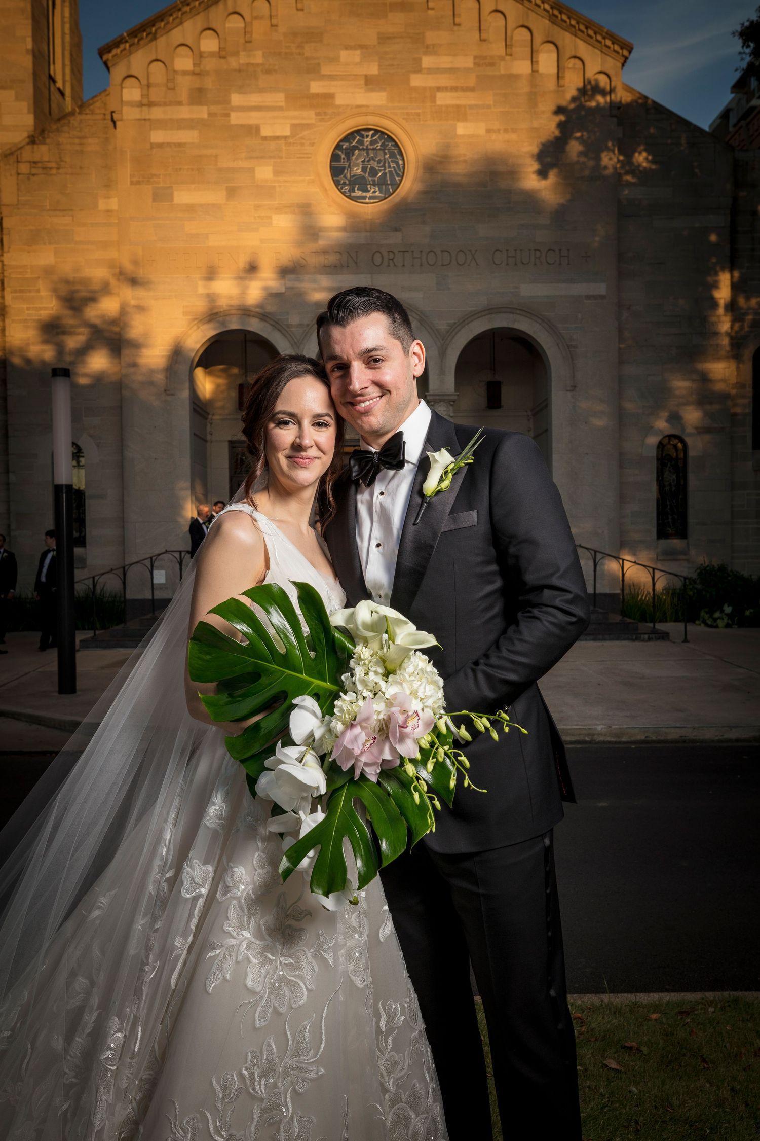 Wedding couple poses outside a stone church facade lit by warm evening light.