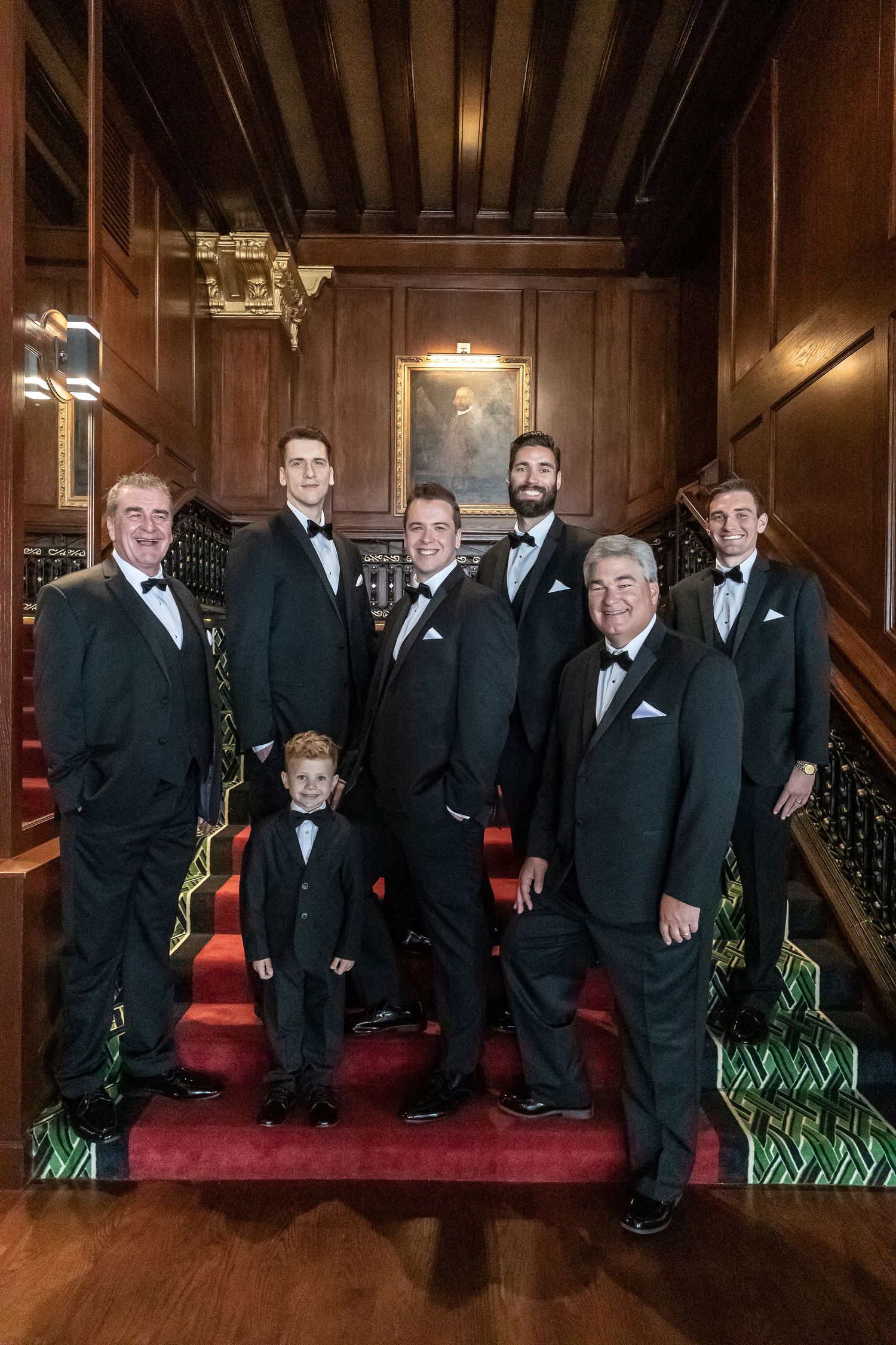 Wedding party in formal black tuxedos poses on red carpeted stairs in an elegant venue.