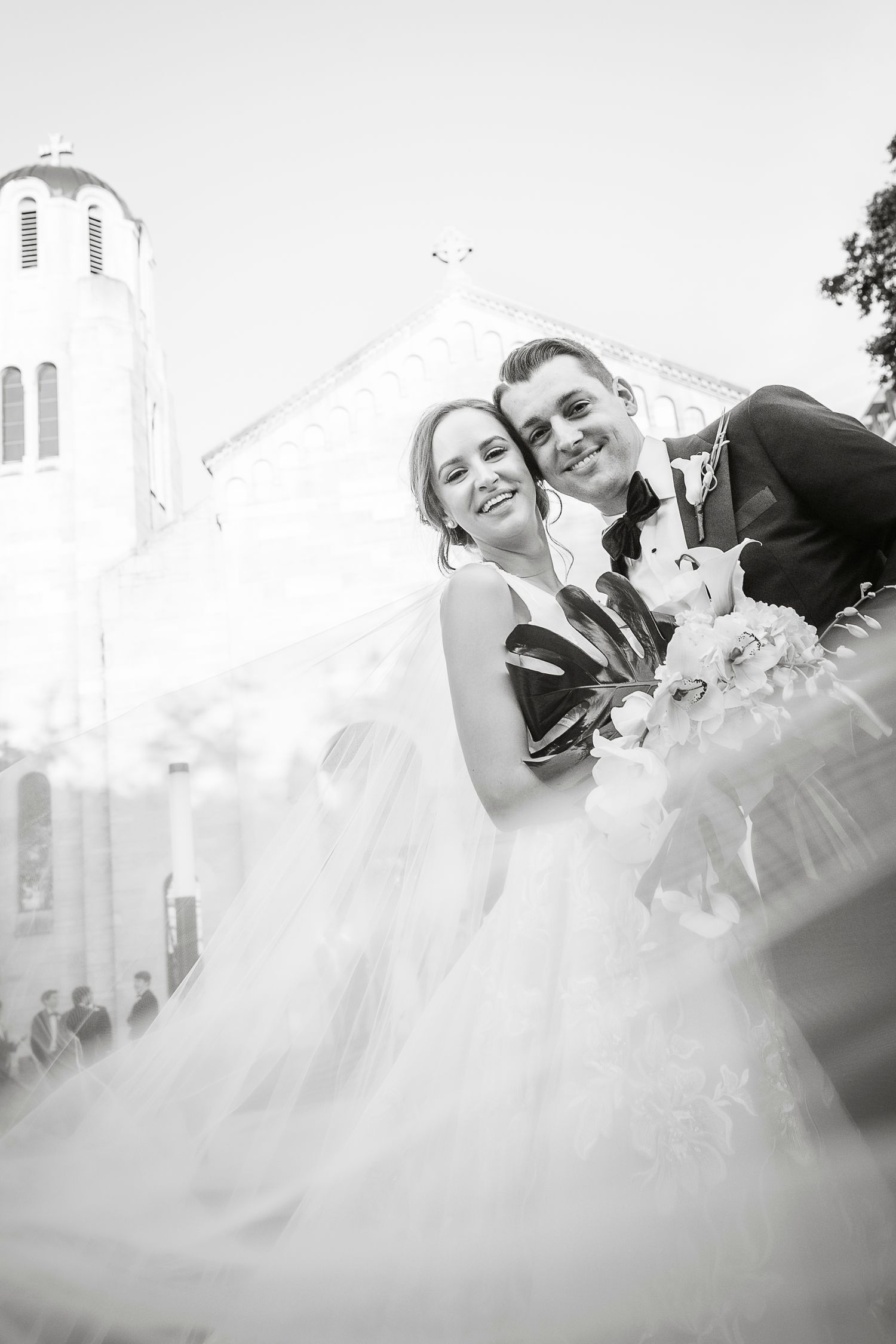 Ethereal black and white wedding photo with flowing veil against a white church backdrop.
