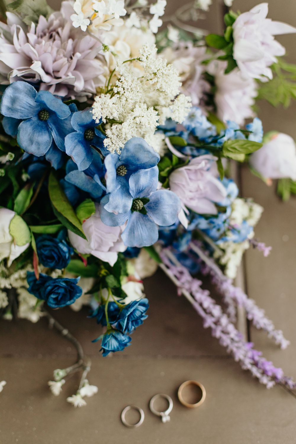 Blue and purple wedding bouquet with rings on neutral background.
