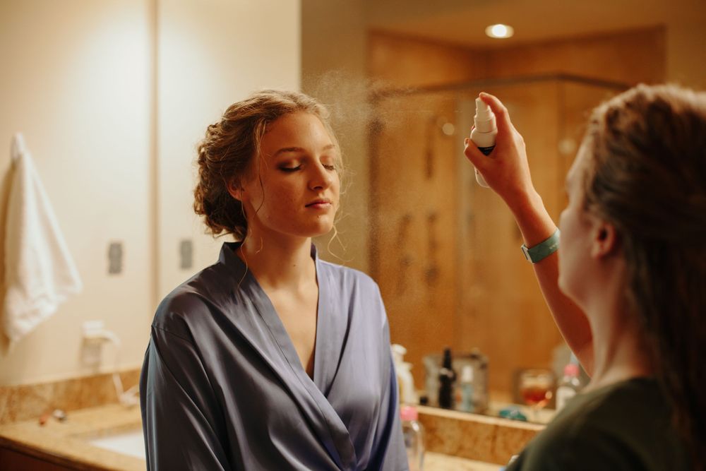 Makeup artist spraying setting spray on bride in blue robe.