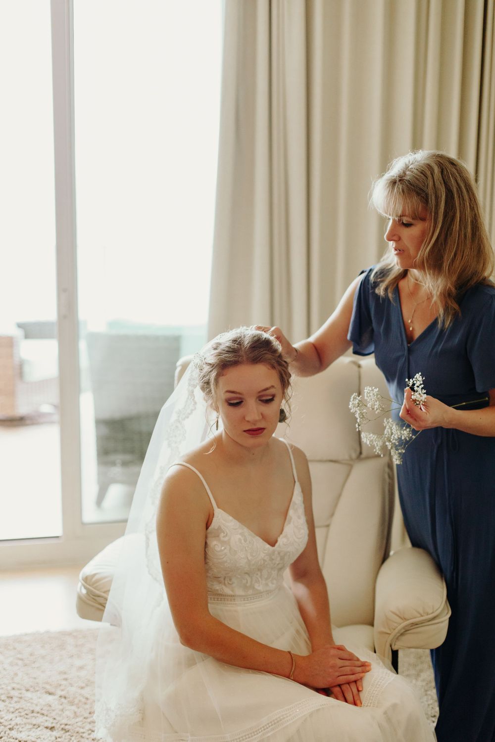 A woman in blue adjusts the bride's veil while she sits in a white chair holding her bouquet.