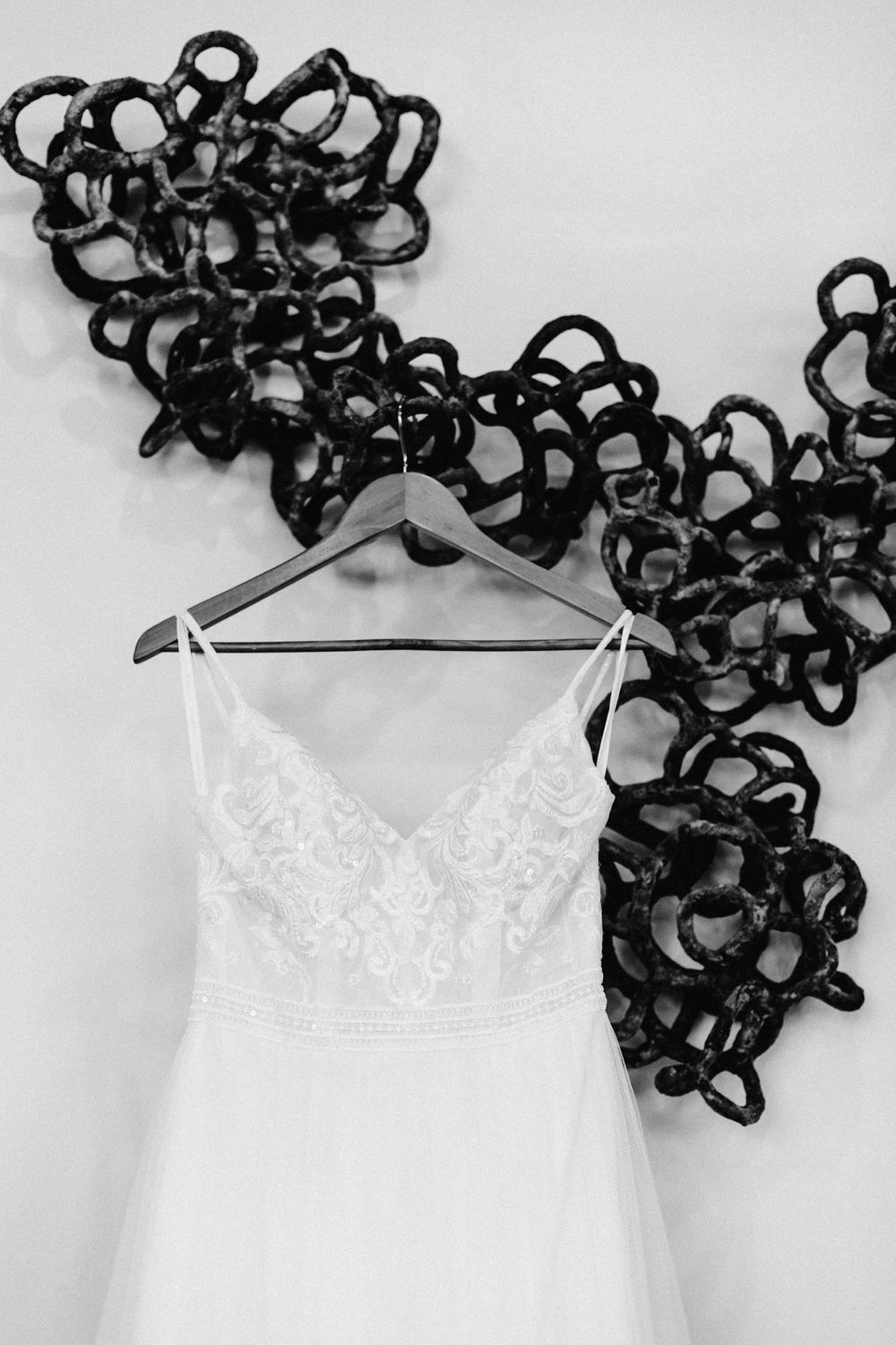 Black and white photo of lace wedding dress on hanger against decorative metal wall art.