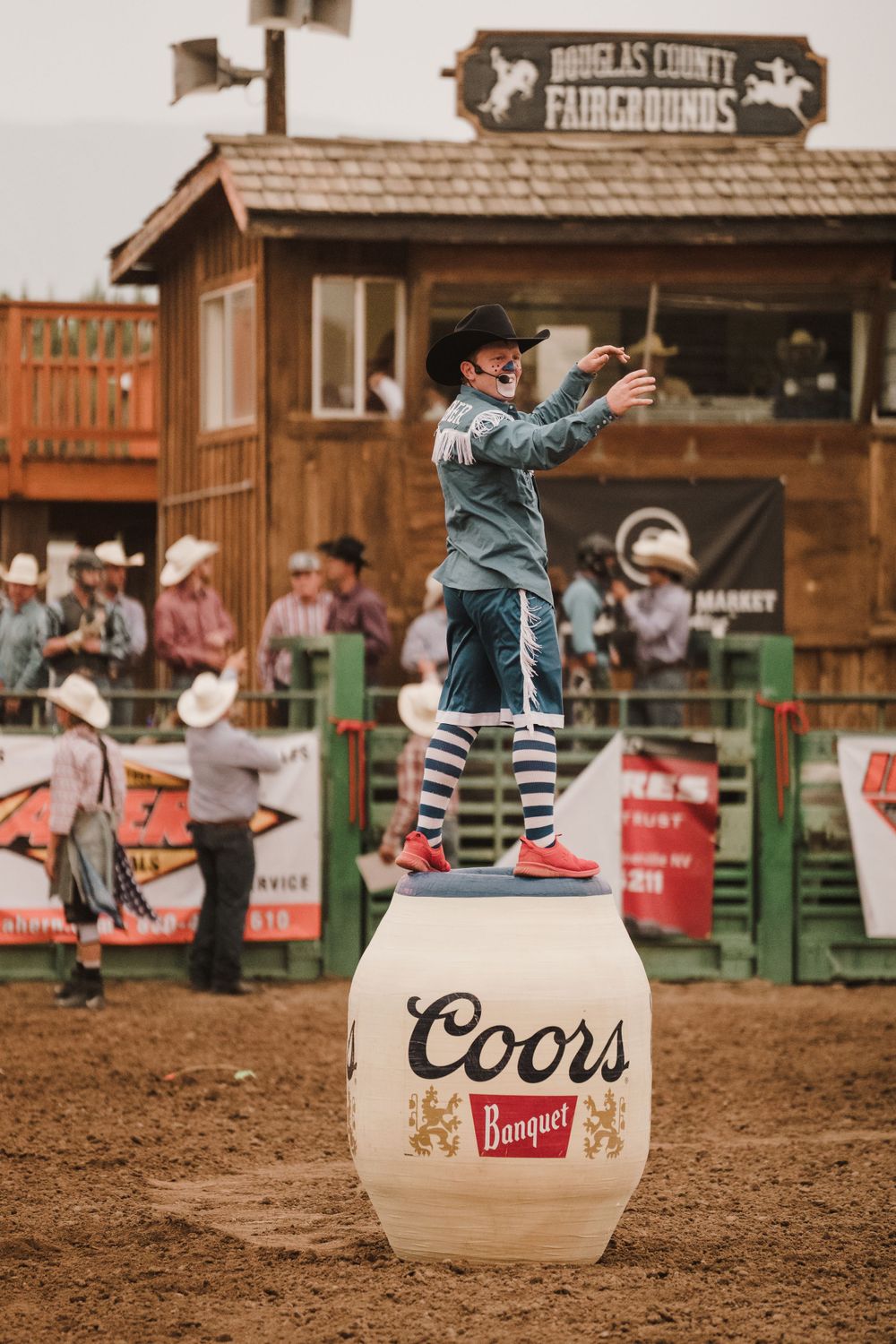 Rodeo performer balancing on large beer barrels in a dirt arena with spectators watching from wooden stands.