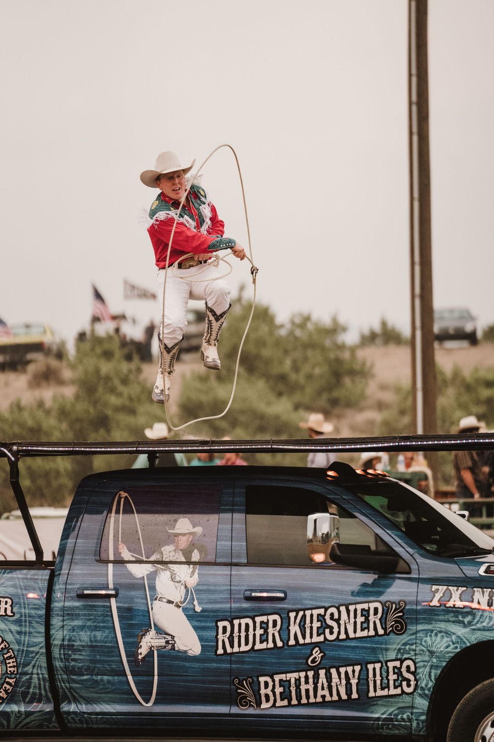 Rodeo performer with lasso jumping over parked cars with names displayed on windows.