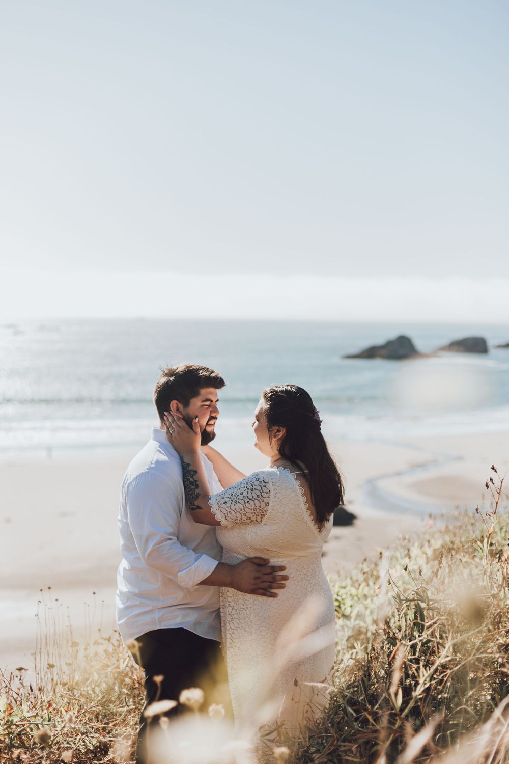 Sun-drenched coastal portrait of an intimate embrace with ocean waves and rocks in the background.
