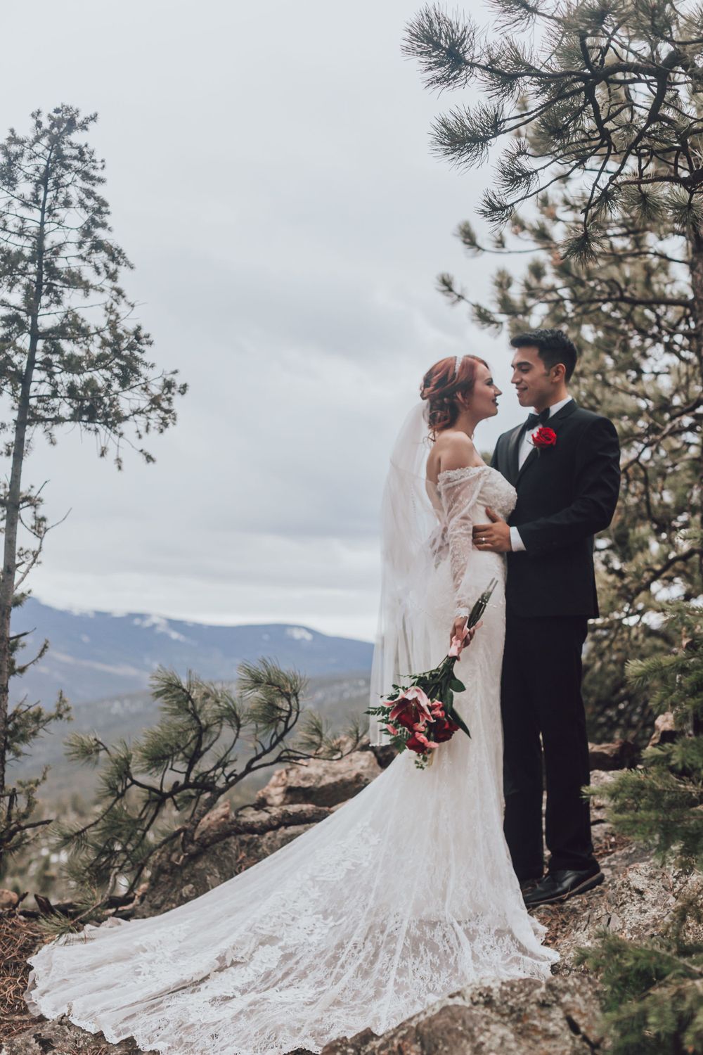 A scenic mountain wedding photo with the couple standing on a rocky outcrop surrounded by pine trees.