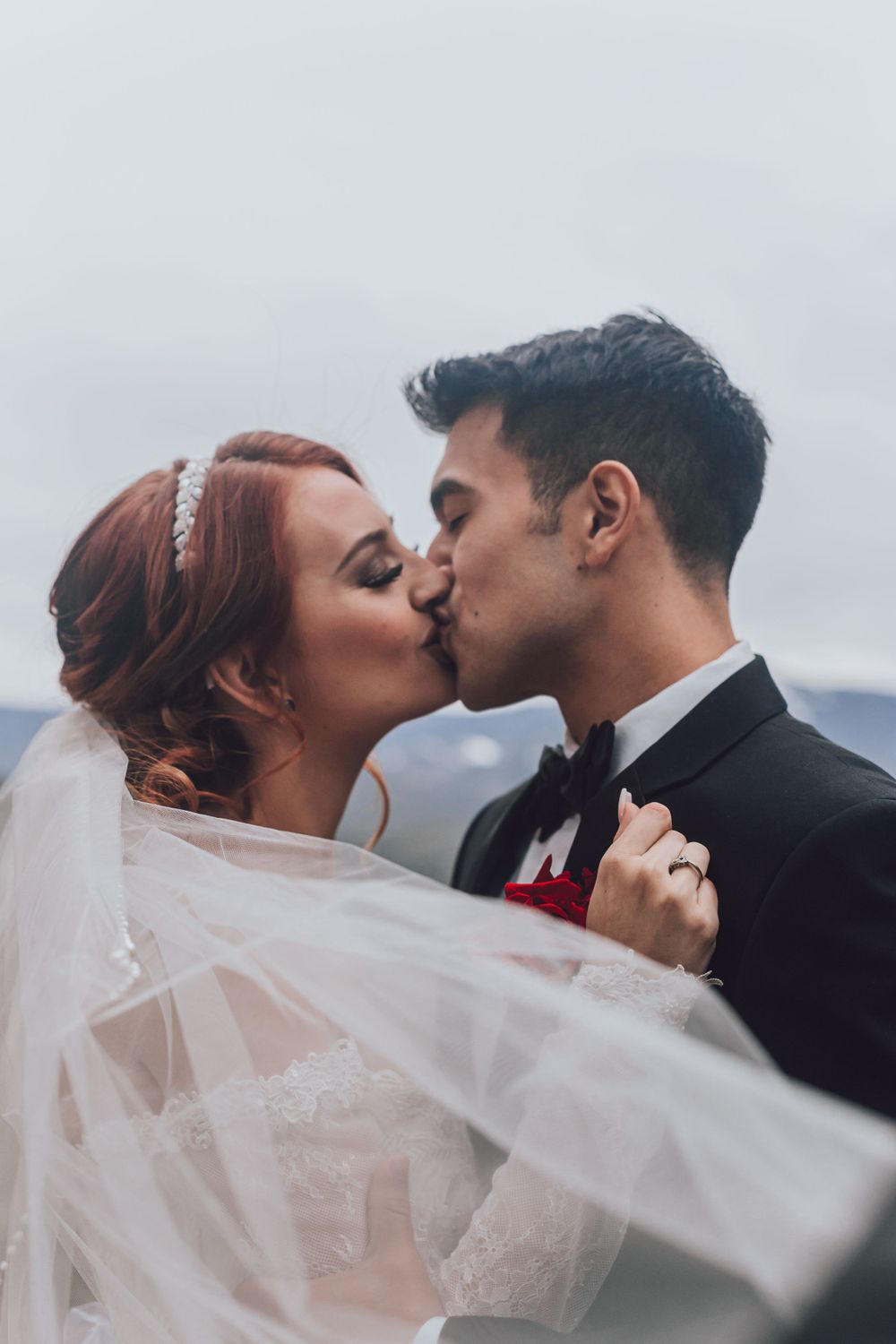 A tender wedding kiss between newlyweds against a cloudy mountain backdrop.