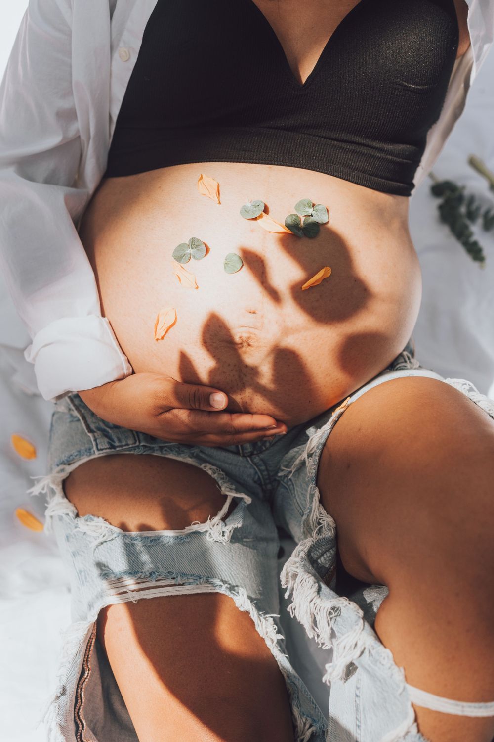 Close-up of pregnant belly in ripped jeans with shadows and petals creating an artistic composition.