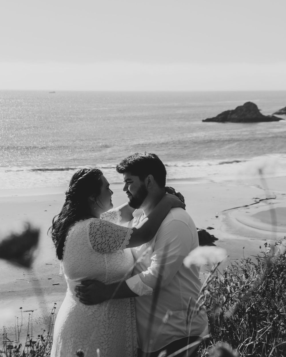 A romantic moment between a couple embracing on a coastal cliff overlooking the ocean in black and white.