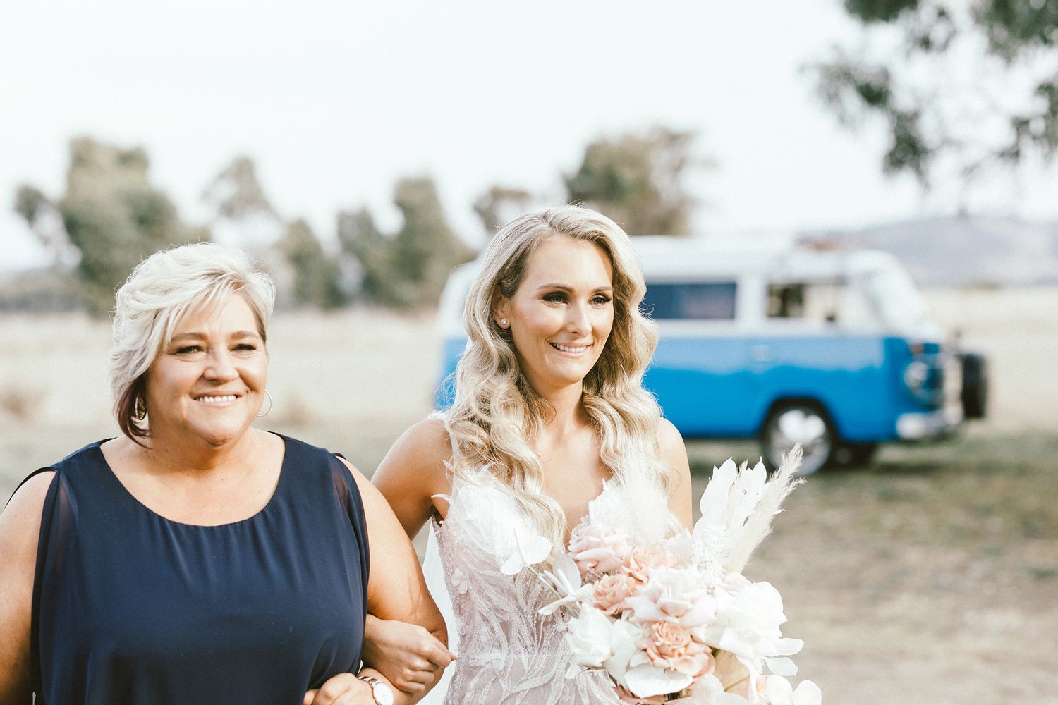 A bride in white lace gown walks with companion through rustic outdoor setting with vintage blue truck in background.