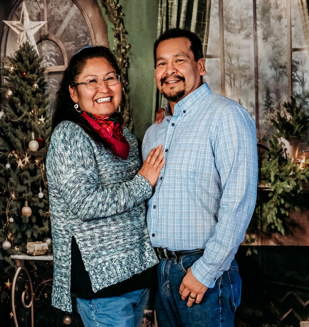 A couple poses together in front of a Christmas backdrop with decorative trees and greenery.