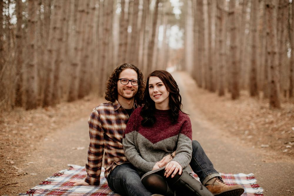 Happy couple sitting on plaid blanket on forest path surrounded by tall trees in autumn.