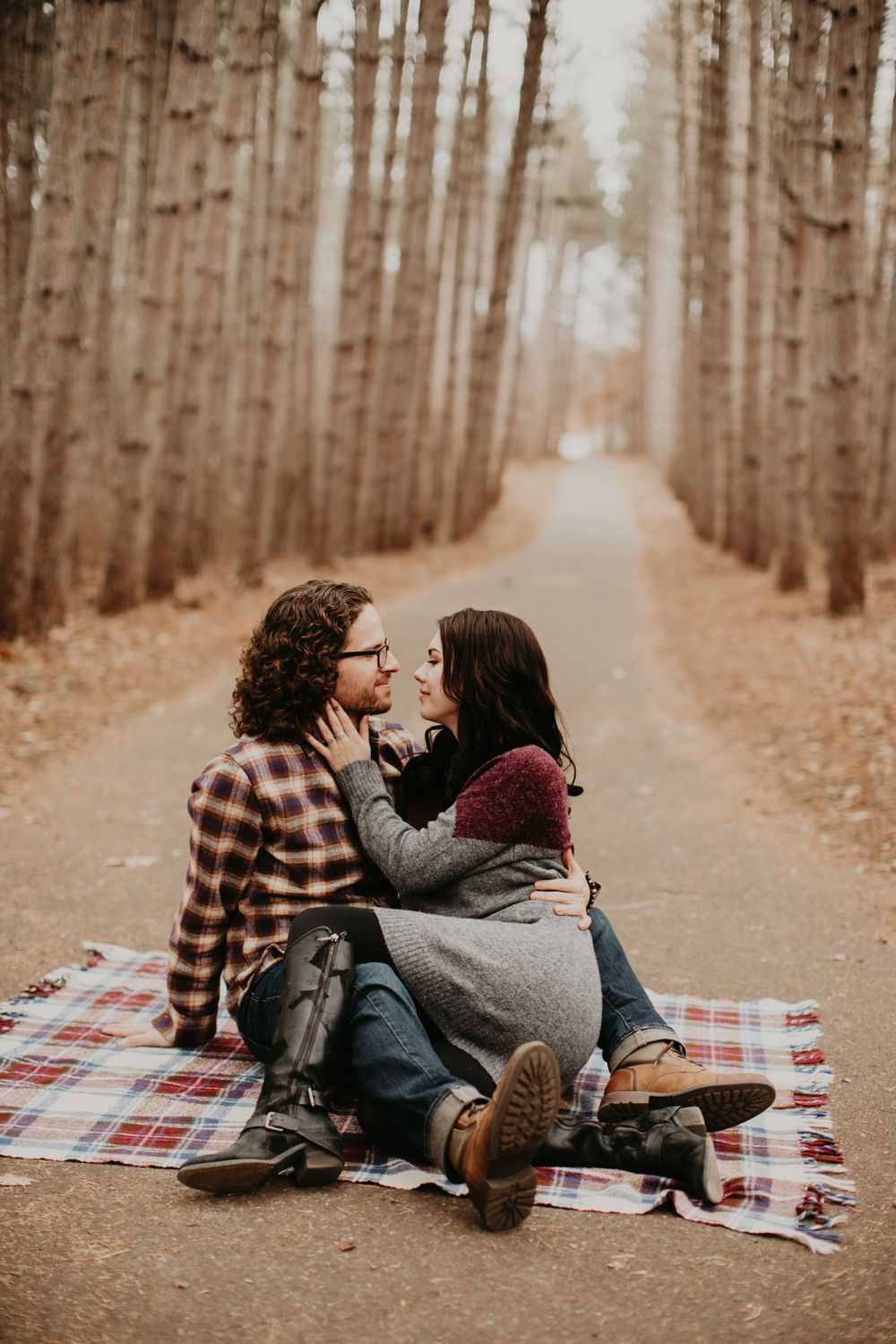 Couple sharing intimate moment on plaid blanket along scenic tree-lined path in fall forest.