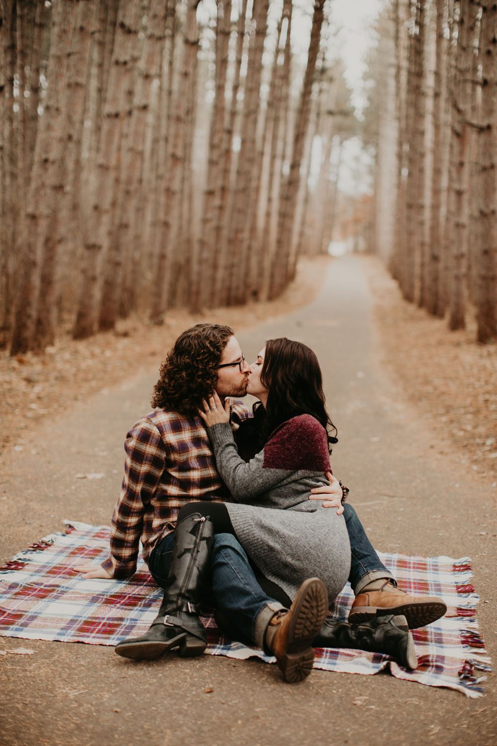 Couple kissing on plaid blanket on a serene tree-lined path in an autumn forest.
