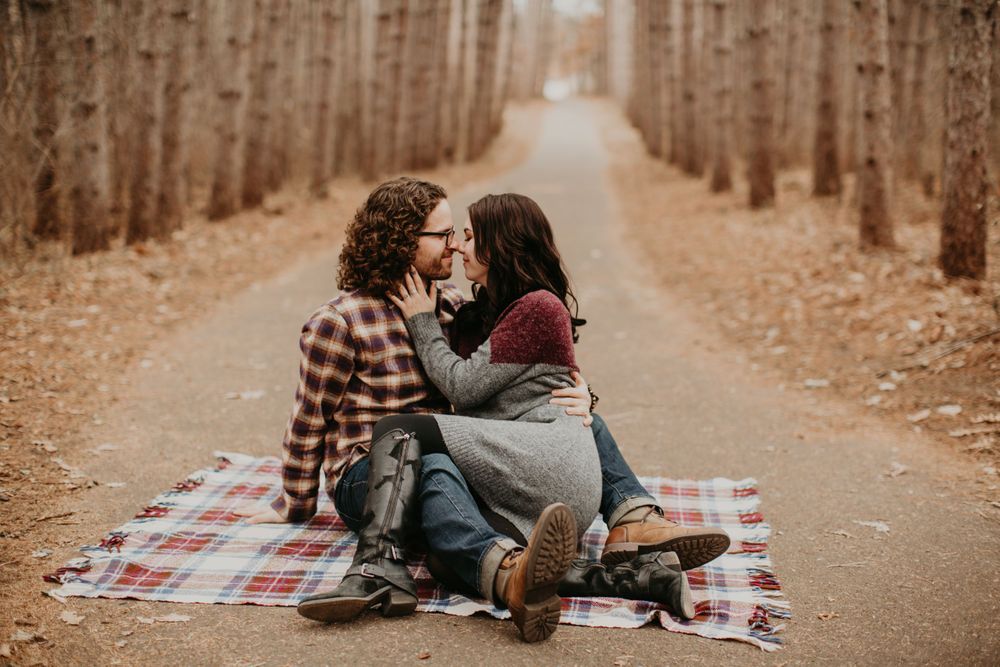 Couple sitting close on plaid blanket, sharing a tender moment on a forest path in fall.