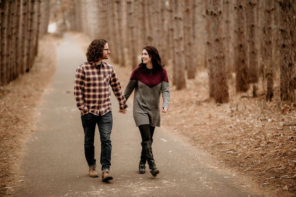 Couple holding hands and smiling while walking along a tree-lined path in autumn.