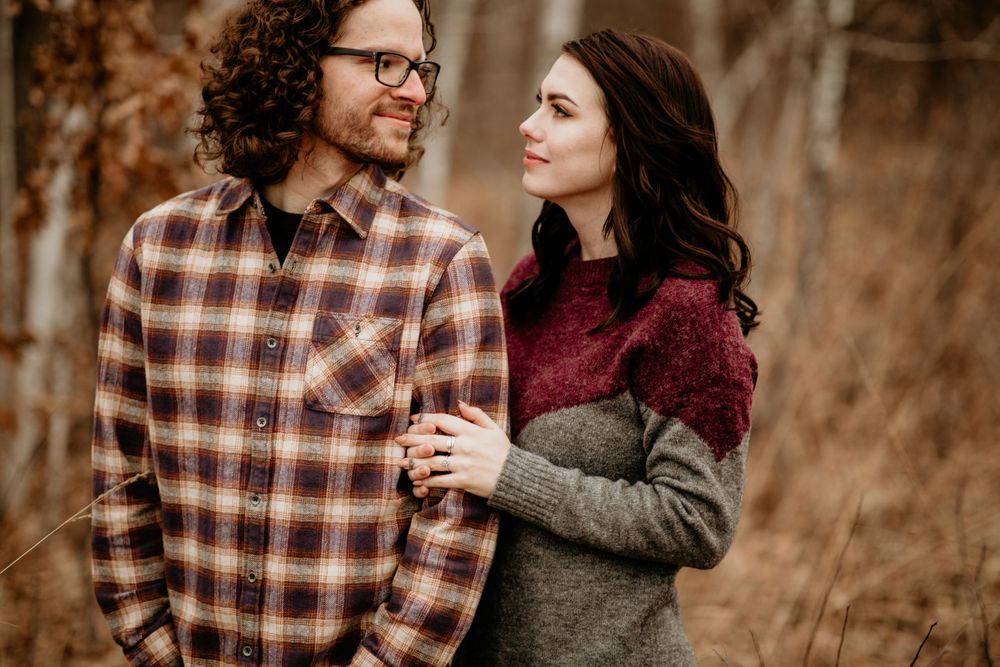 Couple embracing in autumn woods, man in plaid shirt gazing at dark-haired woman in colorblock sweater.