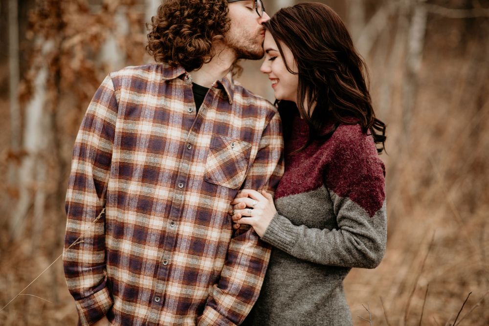 Man kissing woman's forehead tenderly in fall forest, couple wearing cozy flannel and knit sweaters.